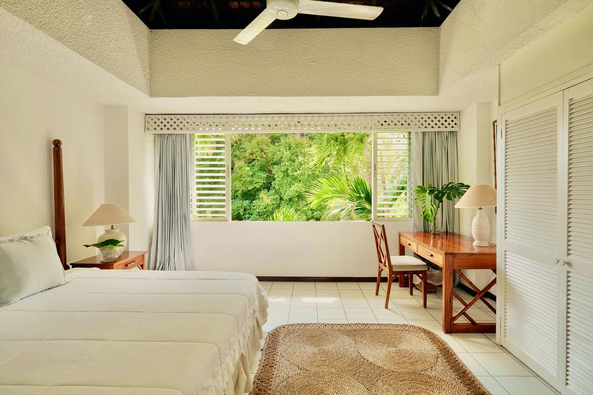 A bedroom with a bed, a desk, a chair, a window with curtains, and a ceiling fan.