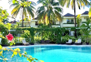 Un área de piscina con sillones de descanso, palmeras y un edificio de fachada blanca.