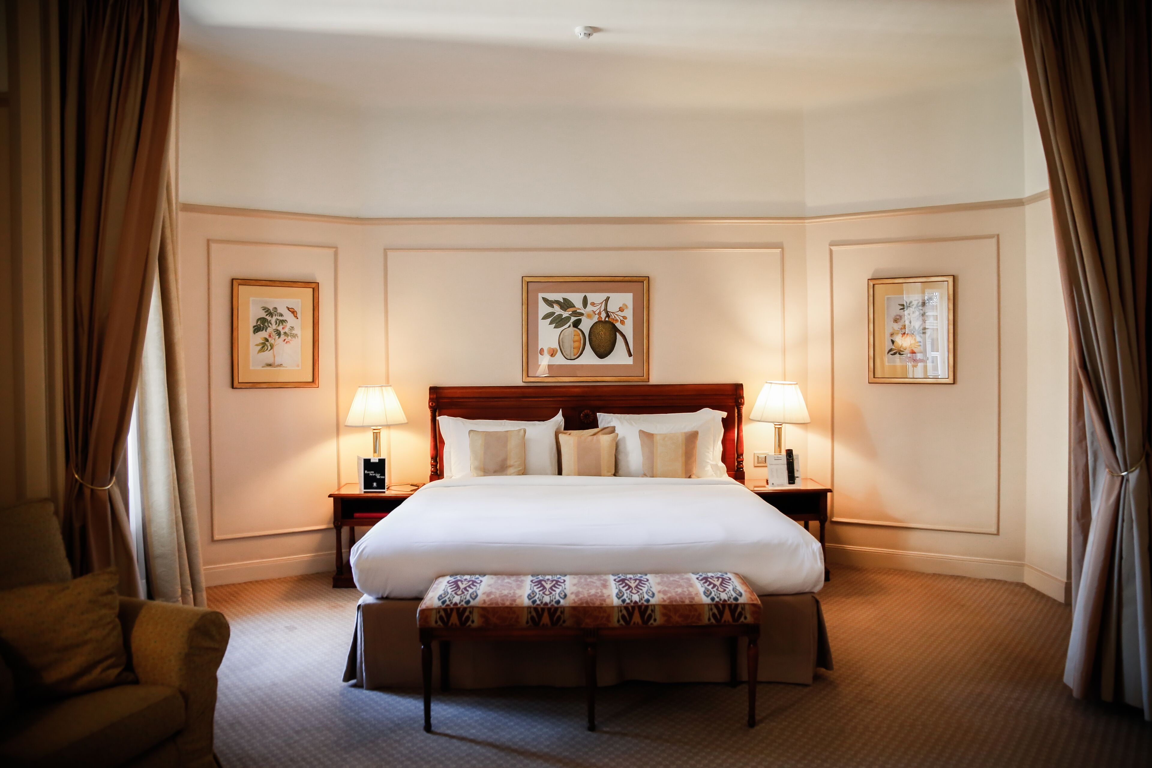 Une chambre d’hôtel avec un grand lit, deux tables de chevet avec des lampes, un banc et des œuvres d’art encadrées accrochées aux murs.