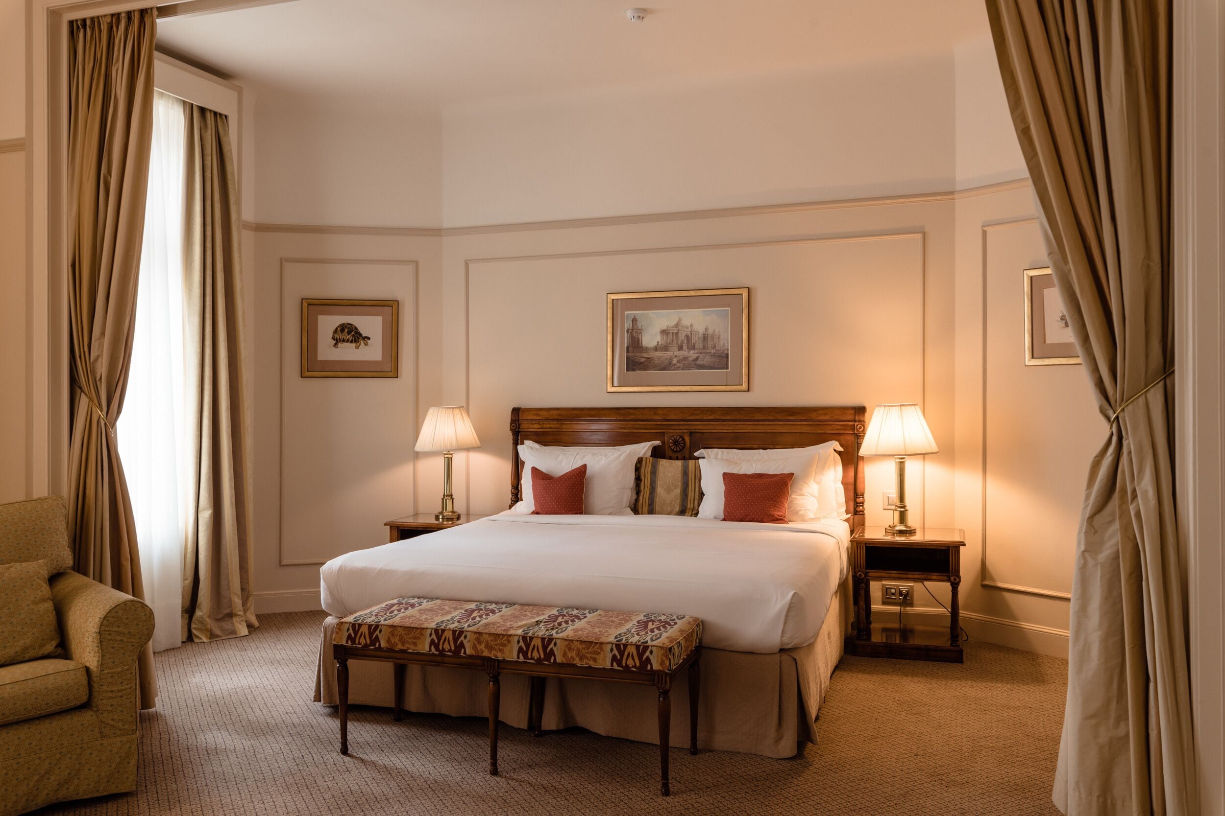 A hotel room with a large bed, two bedside tables with lamps, a bench, a chair, and two framed pictures on the wall.