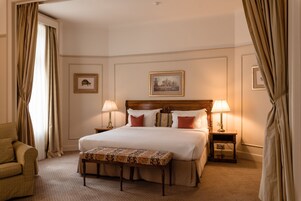 A hotel room with a large bed, two bedside tables with lamps, a bench, a chair, and two framed pictures on the wall.