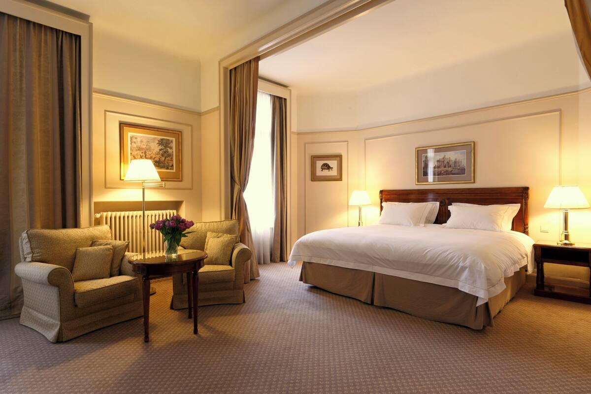 Una habitación de hotel con una cama grande, dos sillones, una mesita con un florero y dos lámparas.