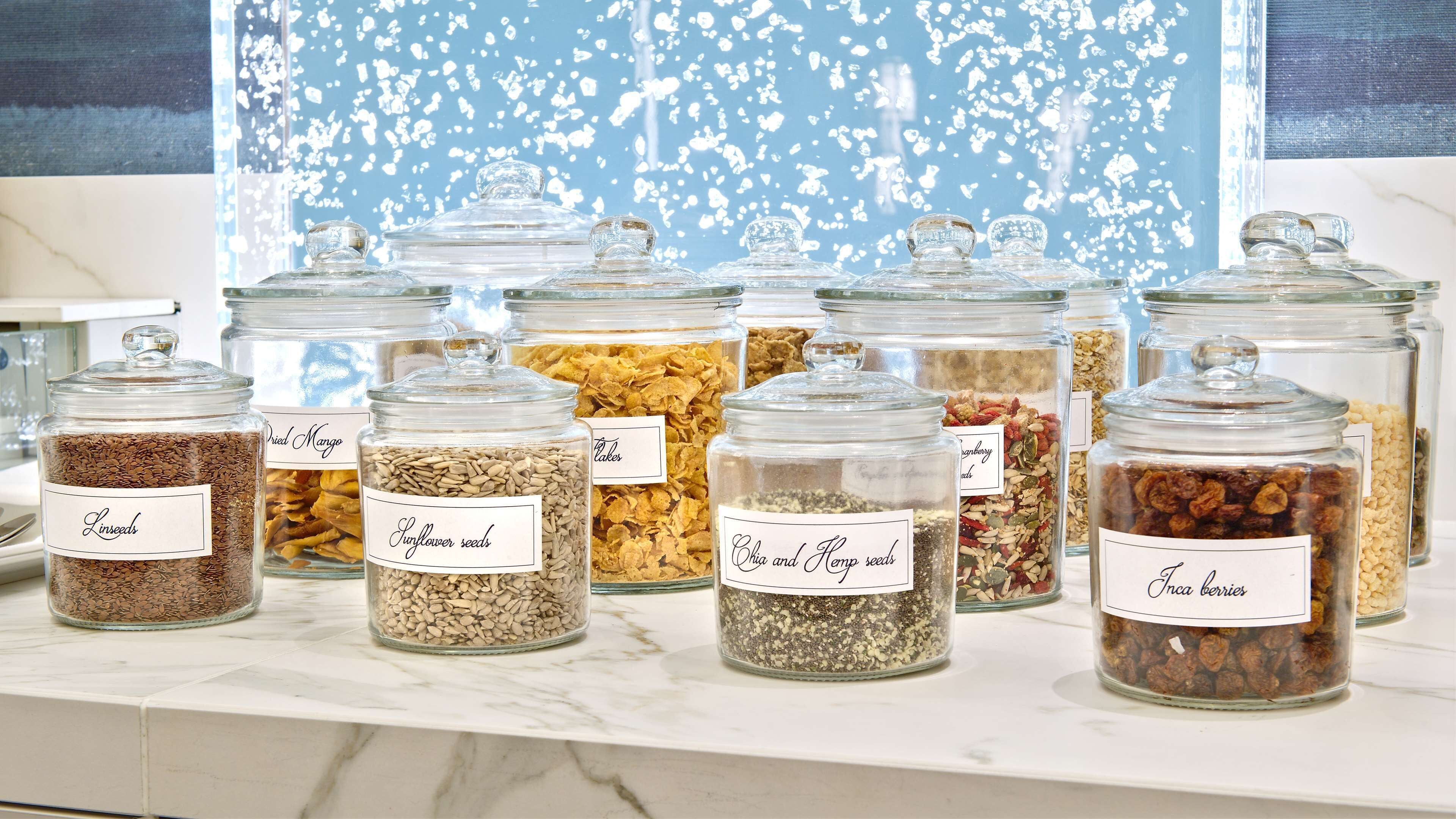 A kitchen counter with multiple glass jars containing various seeds and dried fruits, each labeled with their contents.