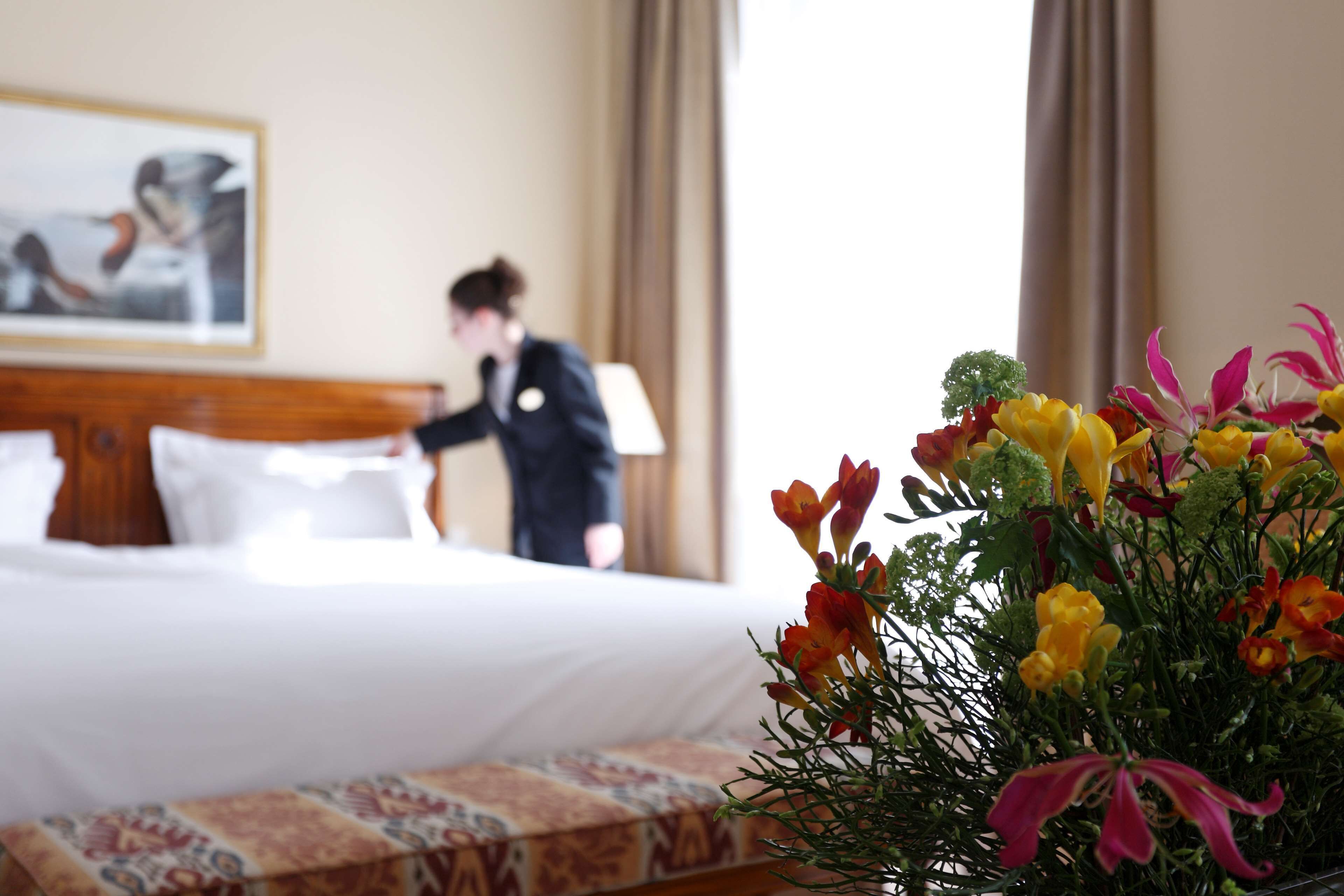 Quarto de hotel com cama bem arrumada, um arranjo floral na mesa de cabeceira e um quadro na parede.