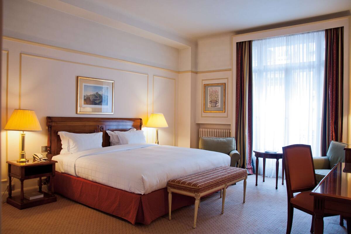 Habitación de hotel con una cama grande, dos lámparas de noche, una mesita de noche de madera, un banco, una mesa de comedor con sillas y vistas al exterior a través de un amplio ventanal con cortinas.