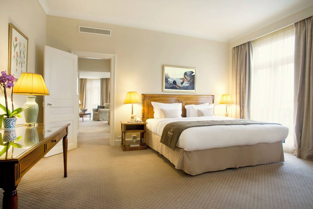 Habitación de hotel con una cama grande, dos lámparas de noche, una mesita de noche de madera, un escritorio con lámpara, un jarrón con flores y un cuadro en la pared.