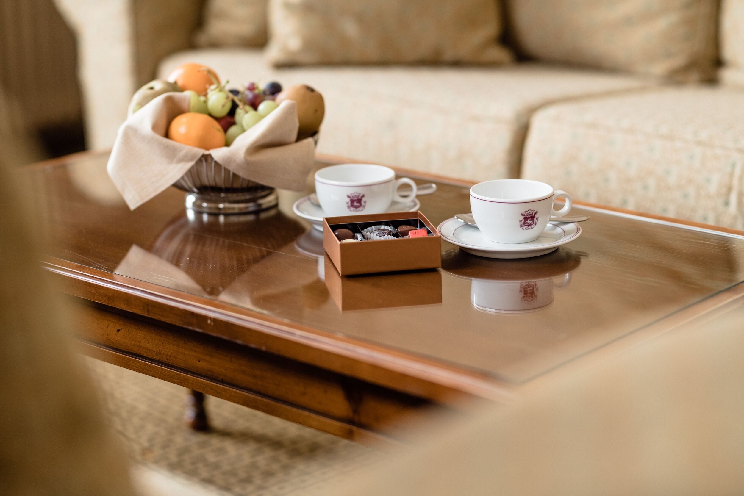 Una mesa de centro de madera con un frutero, dos tazas blancas con platillos y una caja de bombones.