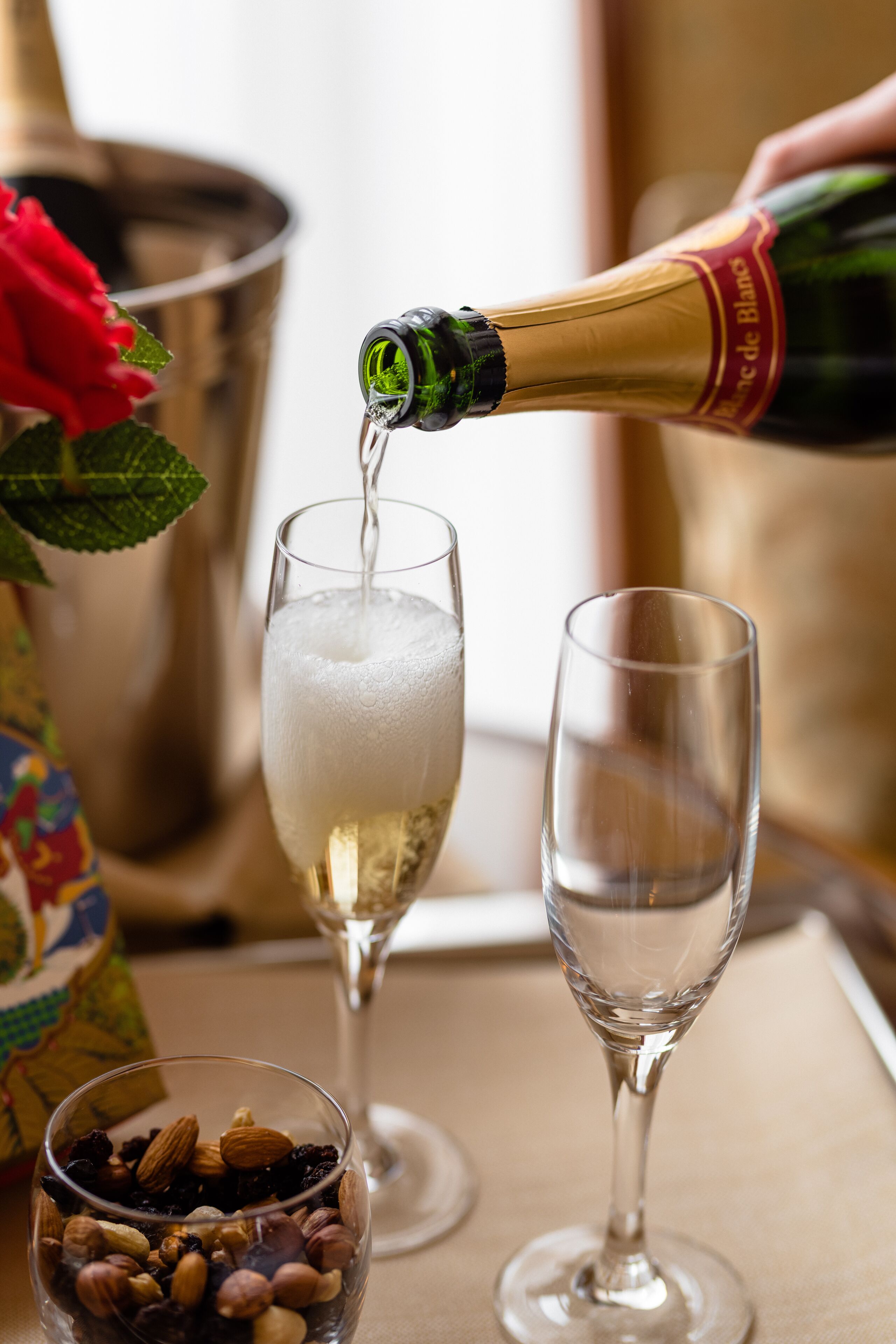 A champagne bottle being poured into a glass, with a bucket and a bowl of nuts in the background.