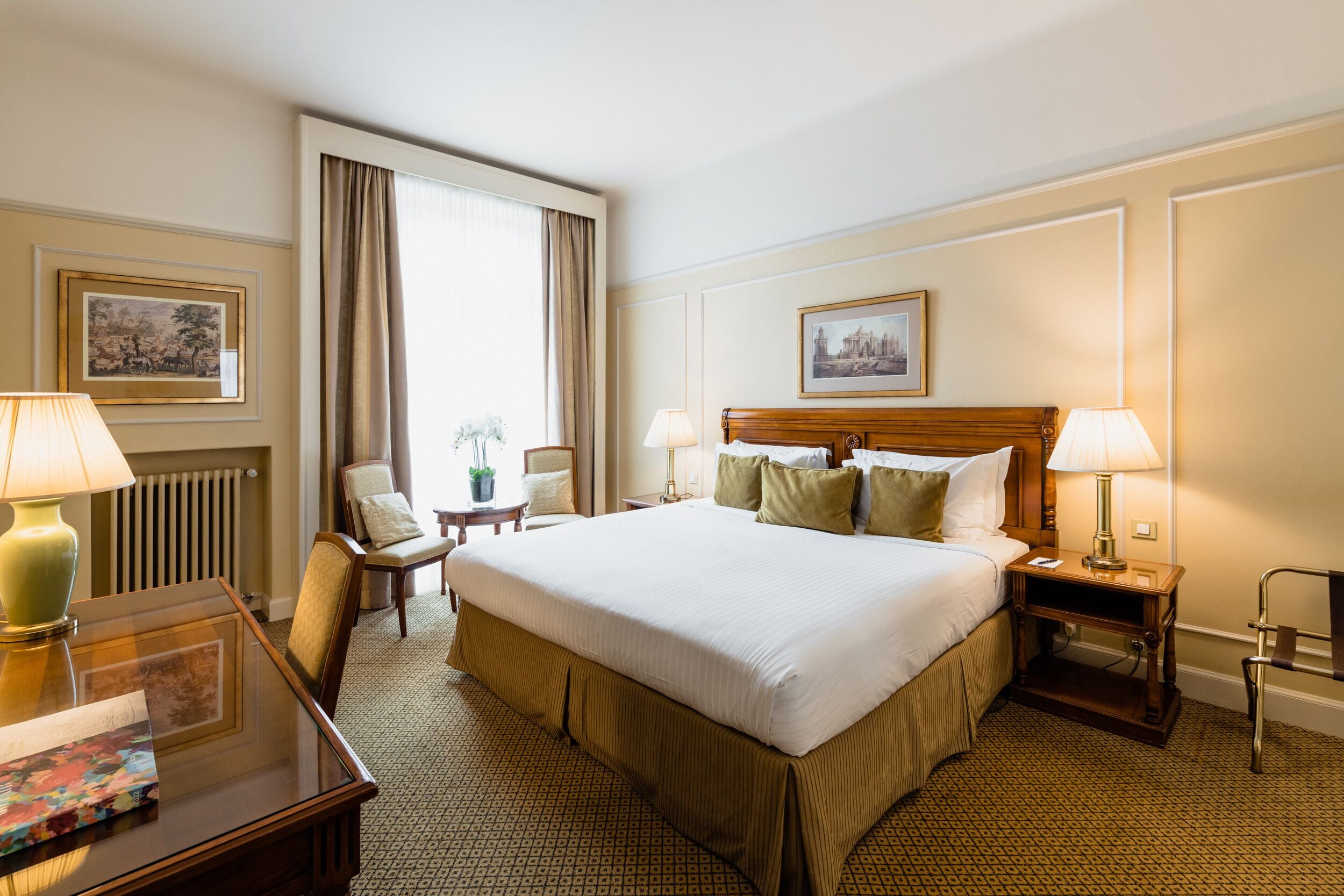 A hotel room with a large bed, two bedside tables with lamps, a chair, a desk with a lamp, and two framed pictures on the walls.