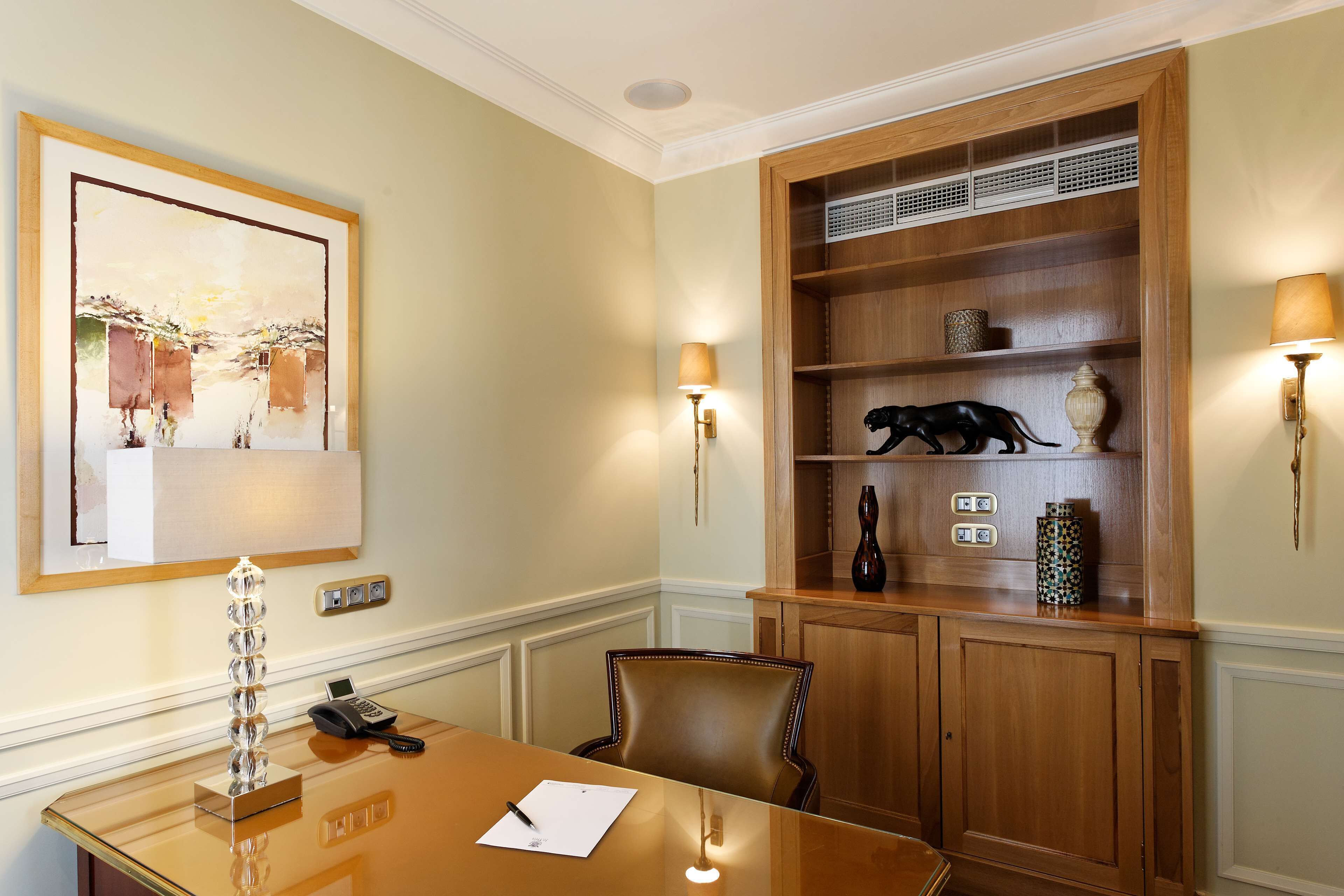 A conference room with a wooden table, a chair, a phone, a lamp, and a shelf with decorative items.