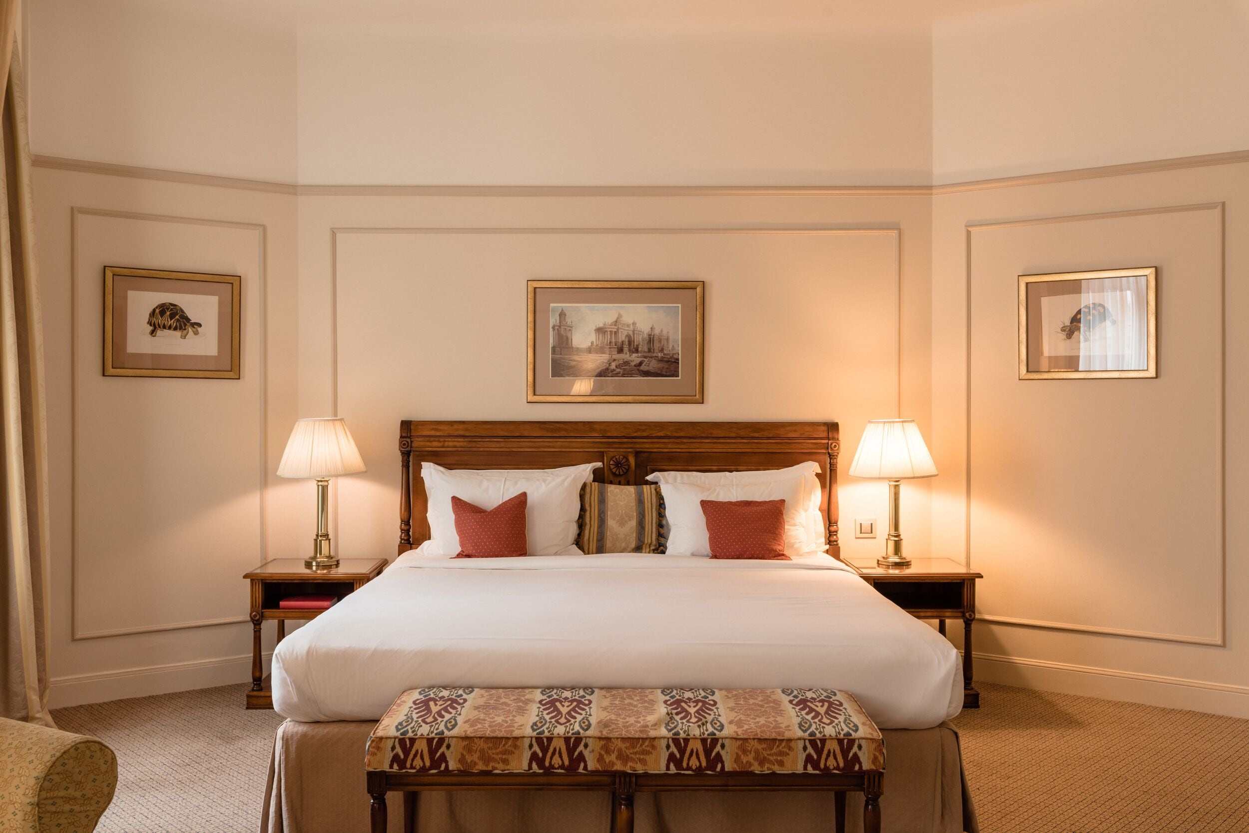 A hotel room with a large bed, two bedside tables, a bench, and framed pictures on the walls.