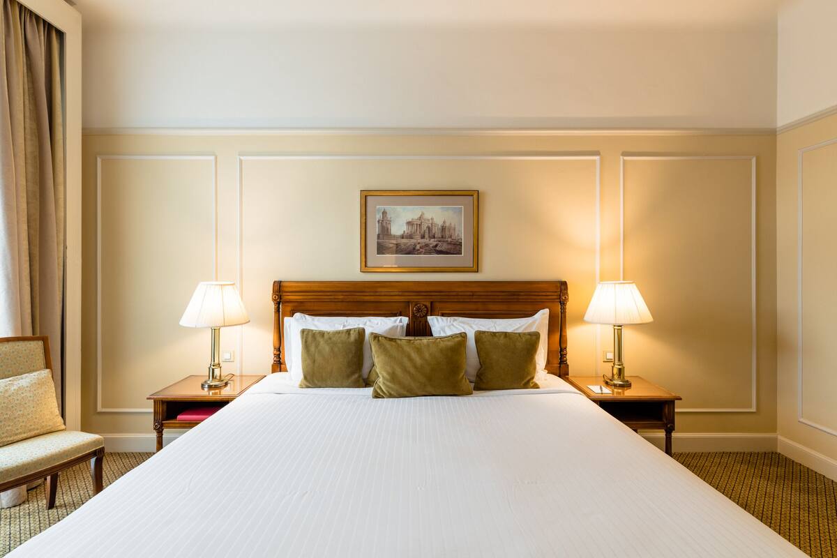Una habitación de hotel con una cama grande, dos mesitas de noche con lámparas, una silla y un cuadro en la pared.