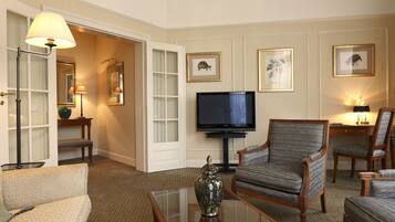 A hotel room with a television, two armchairs, a sofa, a coffee table, and framed artwork on the walls.
