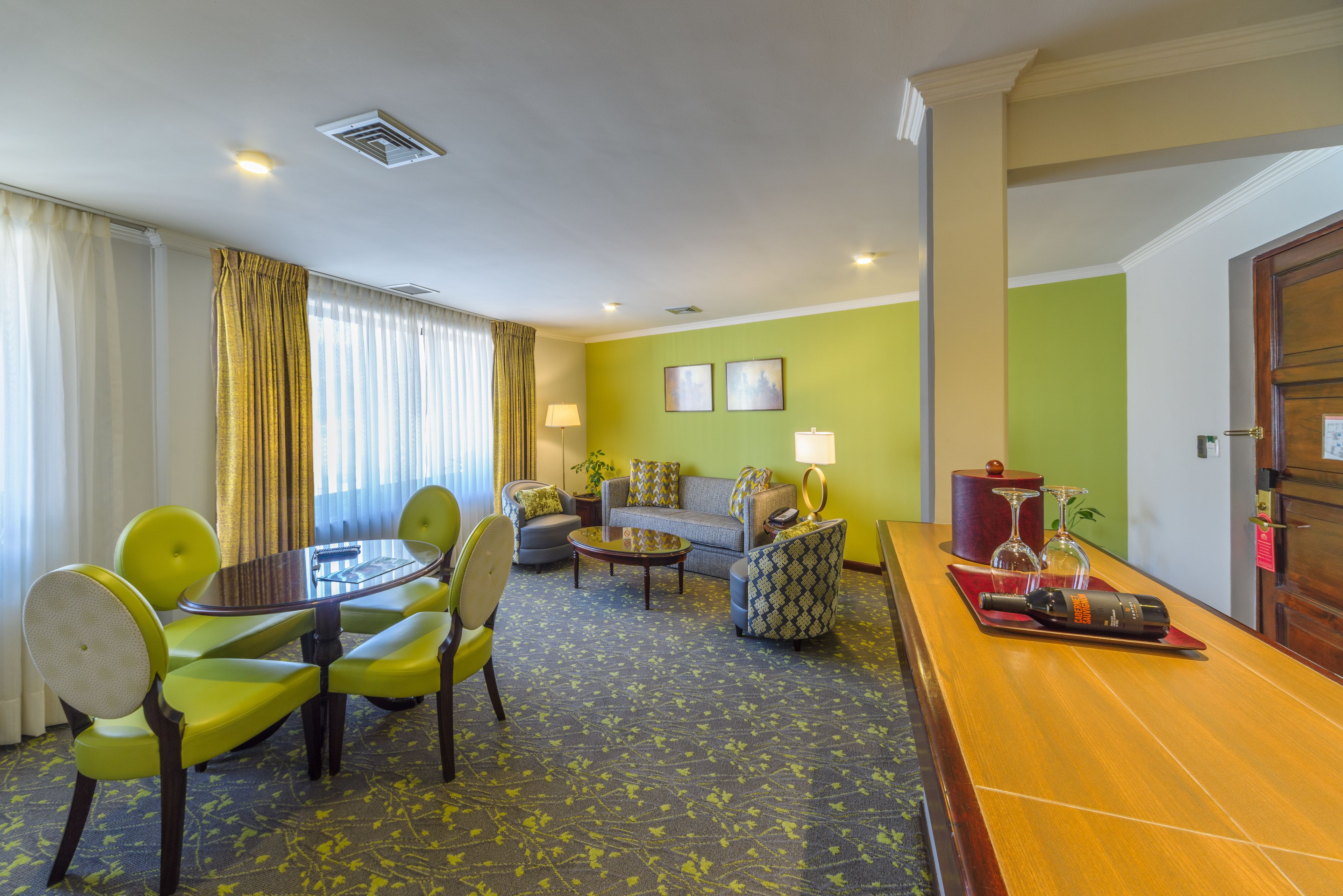 A hotel room with a dining area, a sofa set, a small table with a lamp, and a minibar.