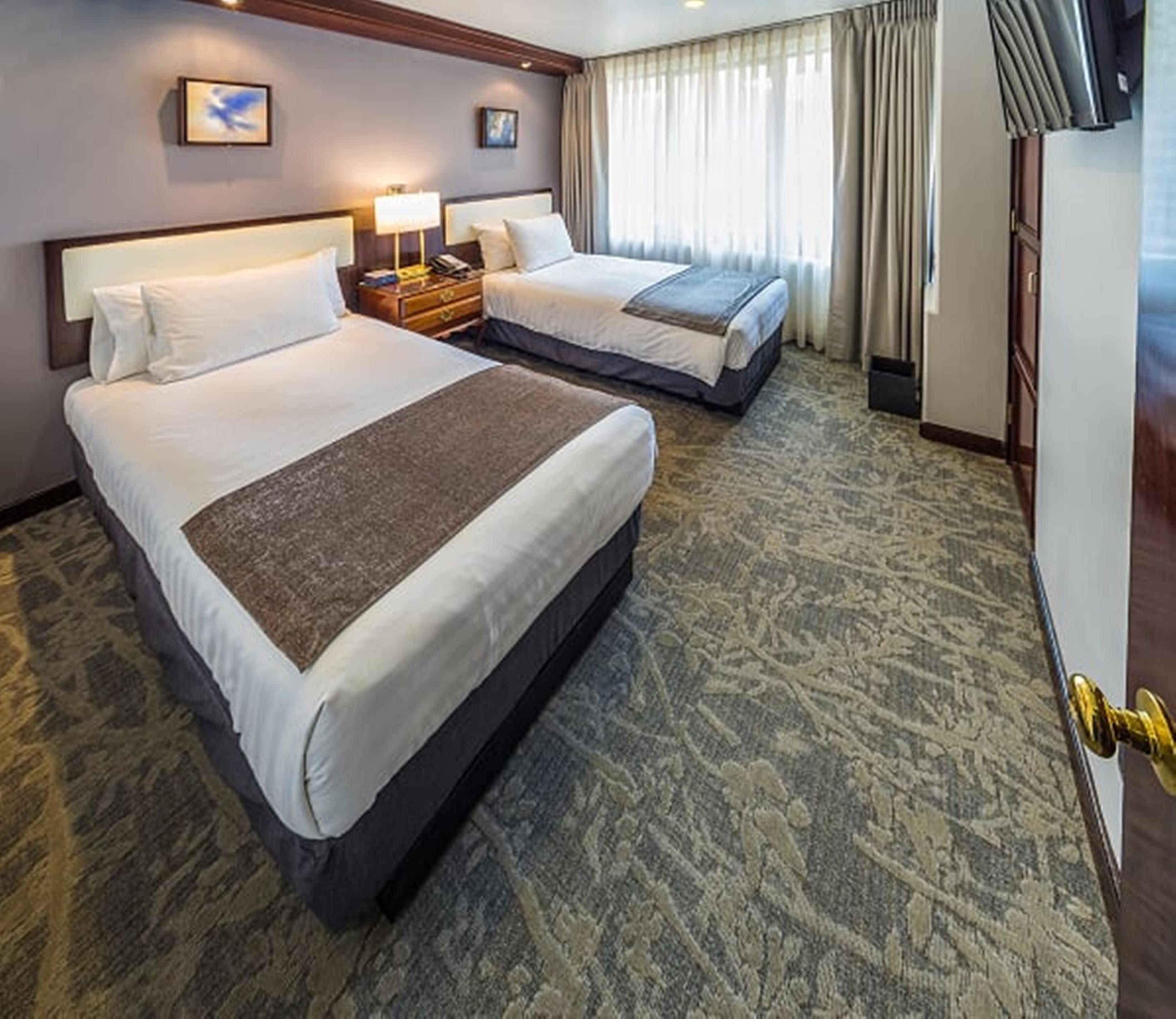 A hotel room with two beds, a carpeted floor, a nightstand with a lamp, and a window with curtains.