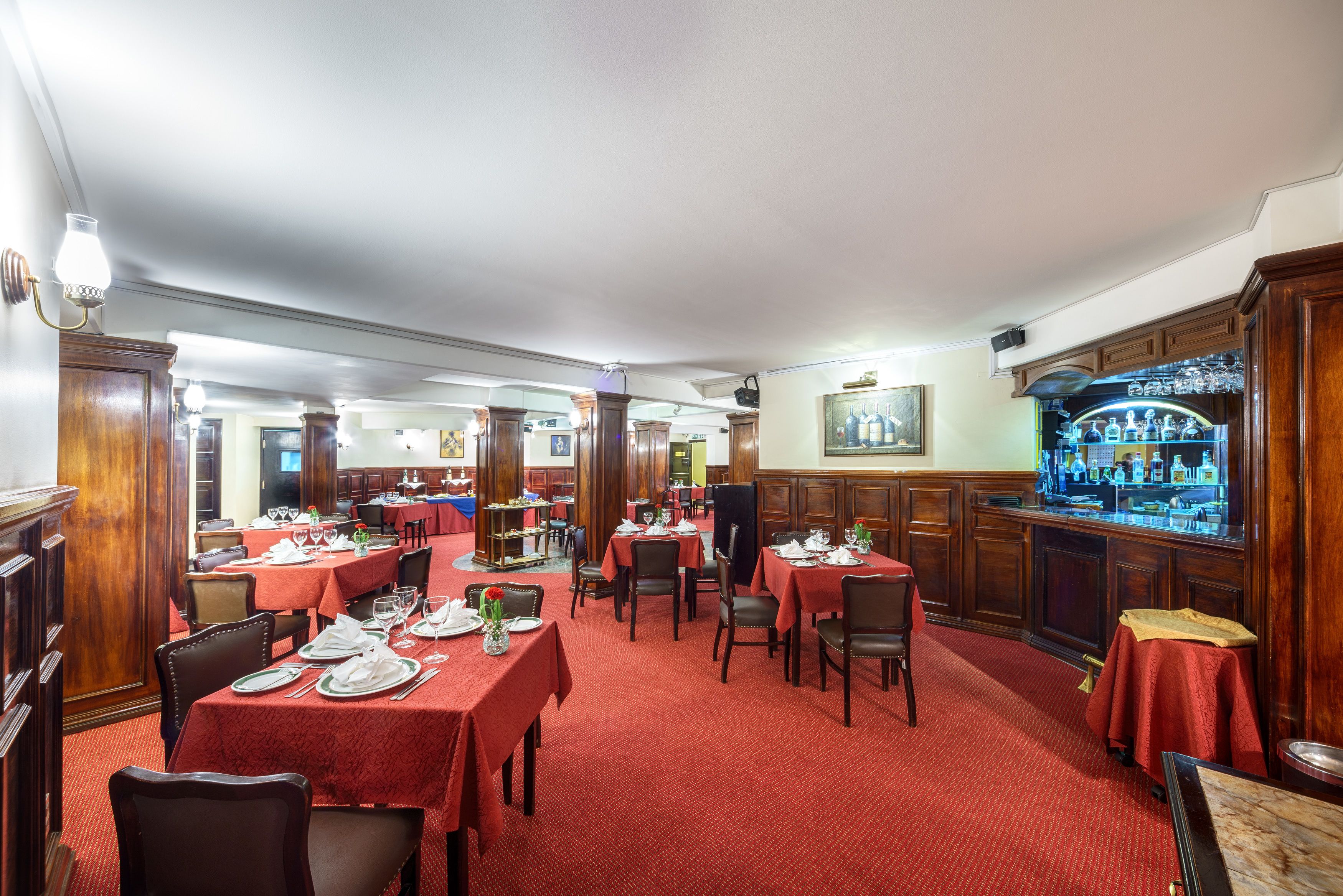 A restaurant with red carpeted tables and chairs, wooden paneling, and a bar area with bottles.