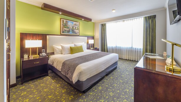 A hotel room with a large bed, bedside lamps, a desk with a telephone, a painting on the wall, and a window with curtains. - Ritz Apart Hotel (La Paz)