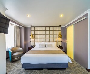 A hotel room with a large bed, two bedside lamps, a curved armchair, and a television mounted on the wall.