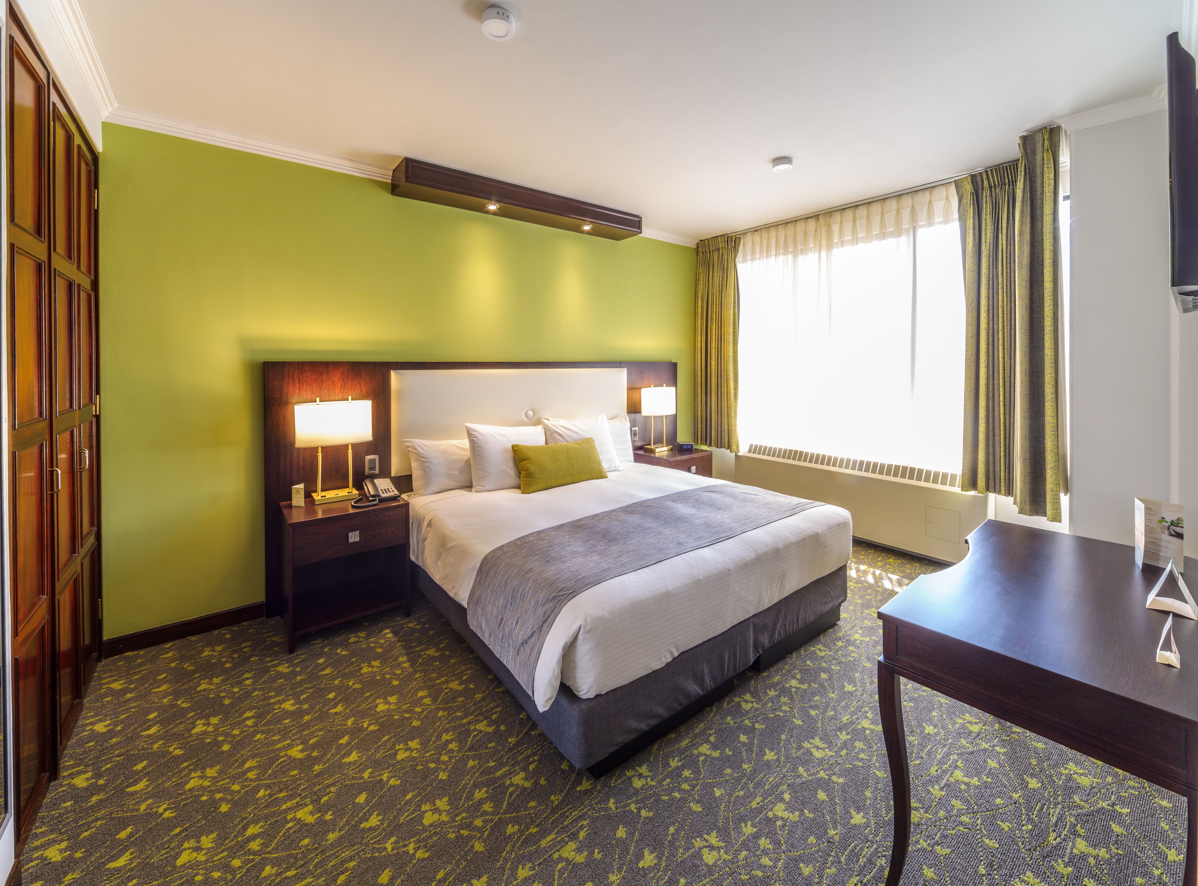 Habitación de hotel con una cama grande, una mesita de noche con lámpara, un buró y un escritorio, además de una ventana con cortinas.