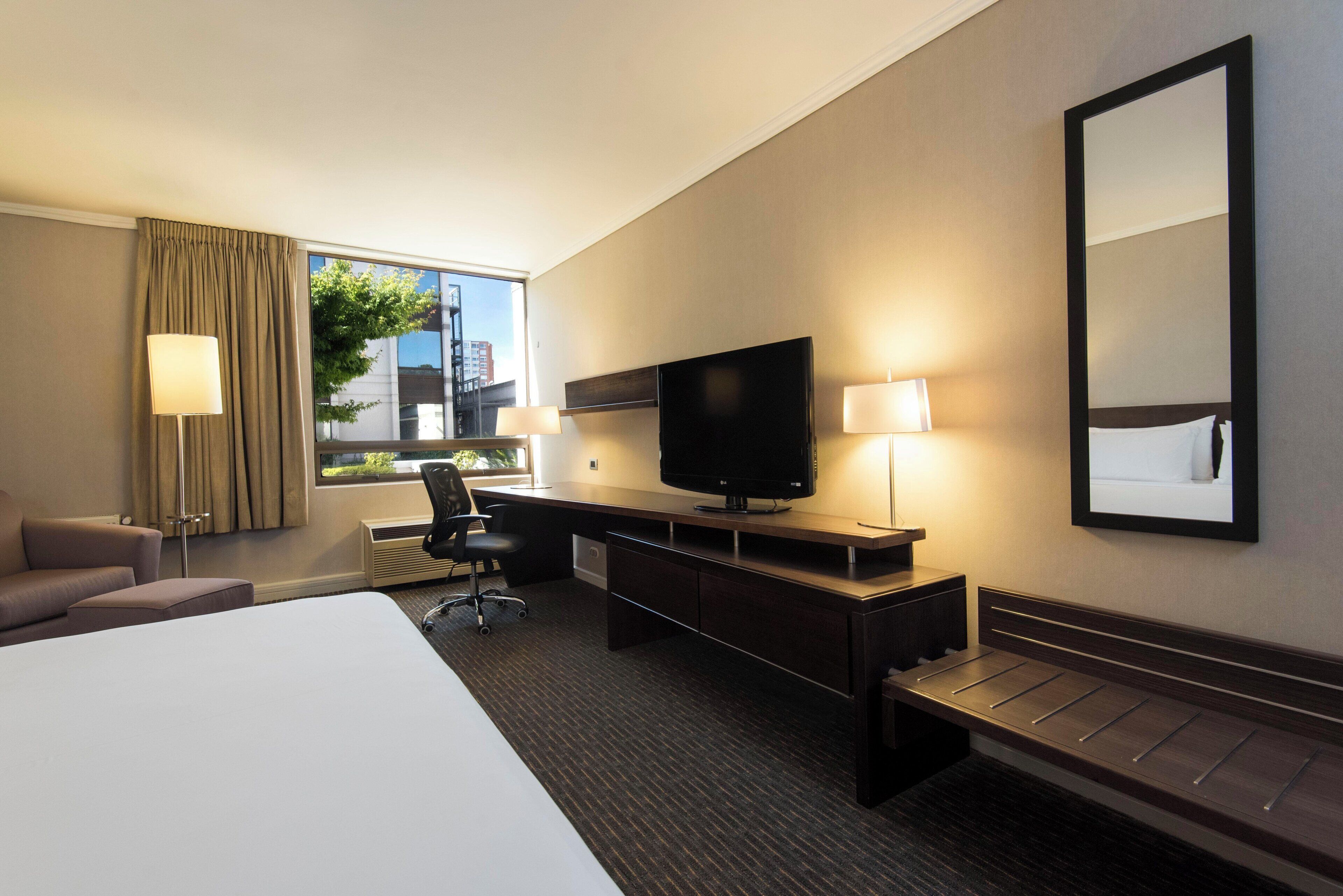Quarto de hotel com cama grande, escrivaninha com televisão, cadeira, abajur e janela com cortinas.