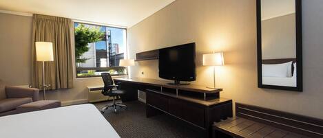 A hotel room with a large bed, a desk with a TV, a chair, a desk lamp, and a window with curtains.