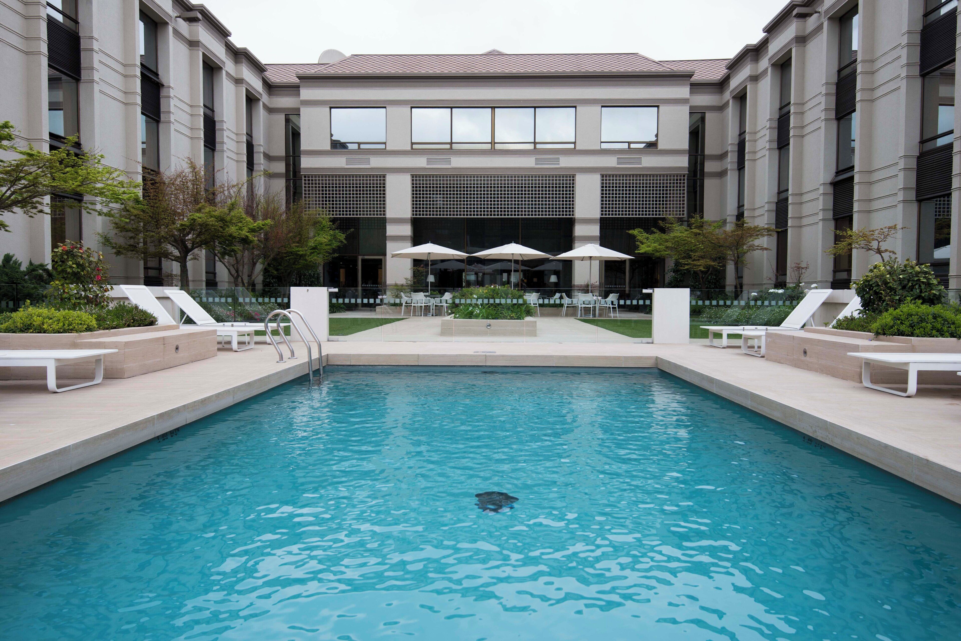 A swimming pool with lounge chairs and umbrellas, surrounded by a building with large windows and a tiled roof.