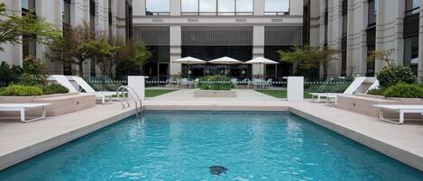 A swimming pool with lounge chairs and umbrellas, surrounded by a building with large windows and a tiled roof.