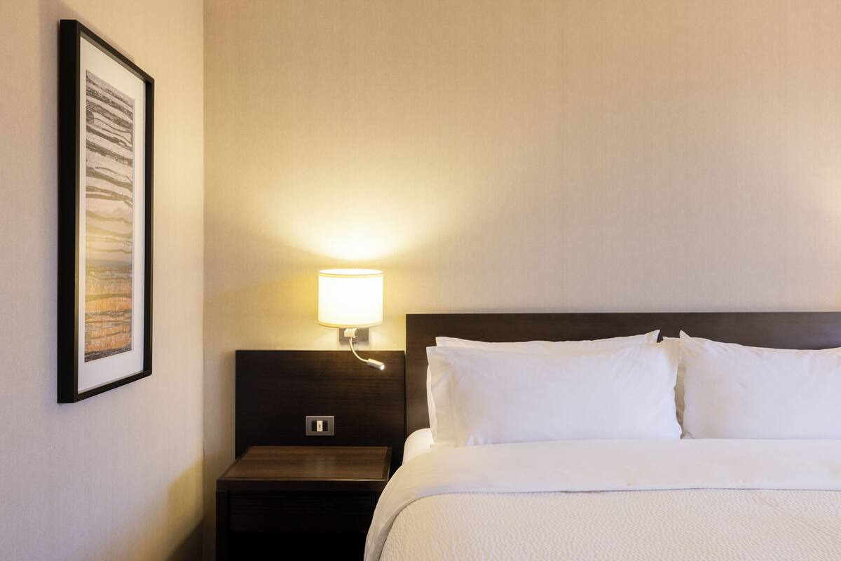A hotel room with a neatly made bed, a bedside table, a wall-mounted lamp, and a framed picture on the wall.
