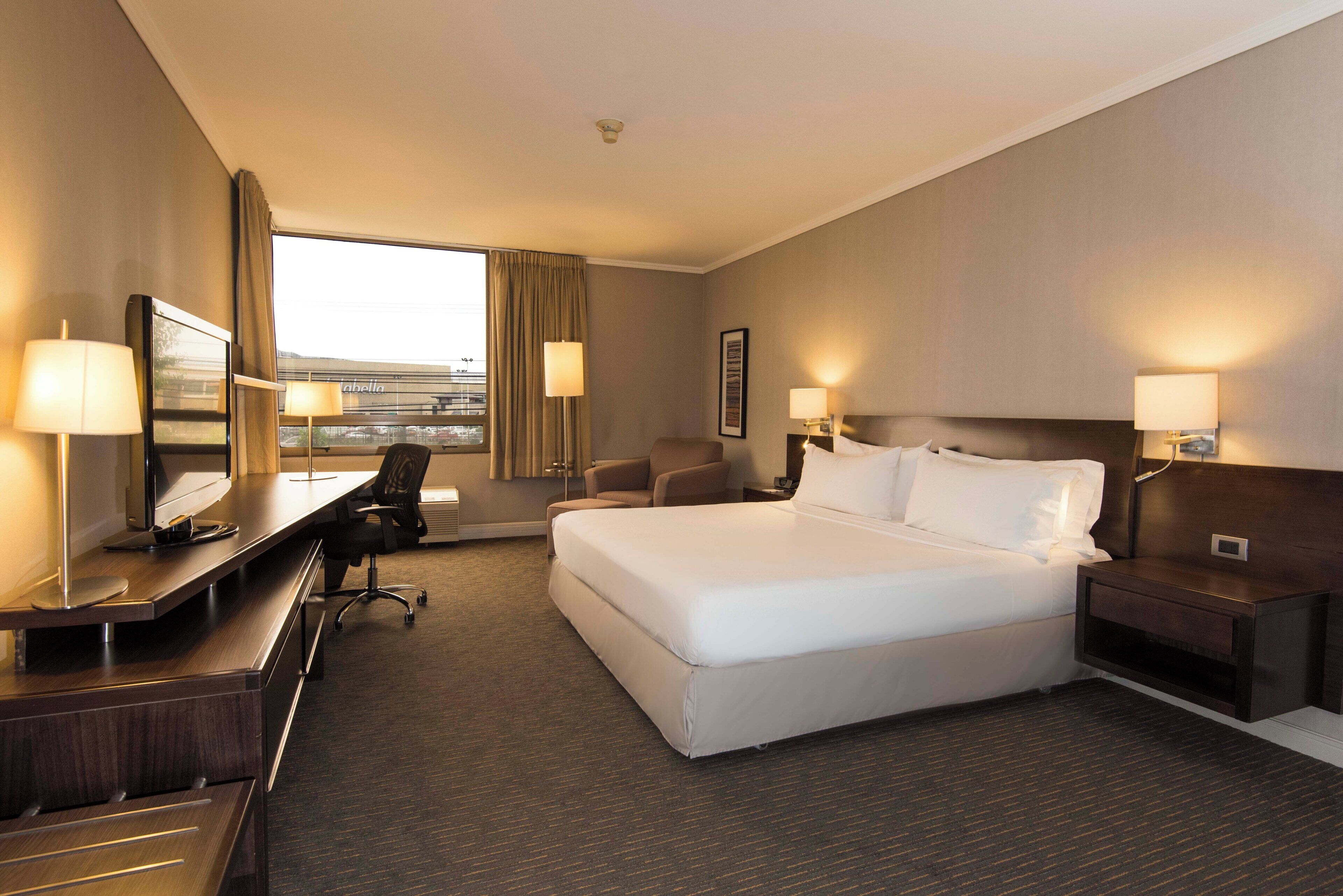 A hotel room with a large bed, a desk with a chair, a TV, and a window with curtains.