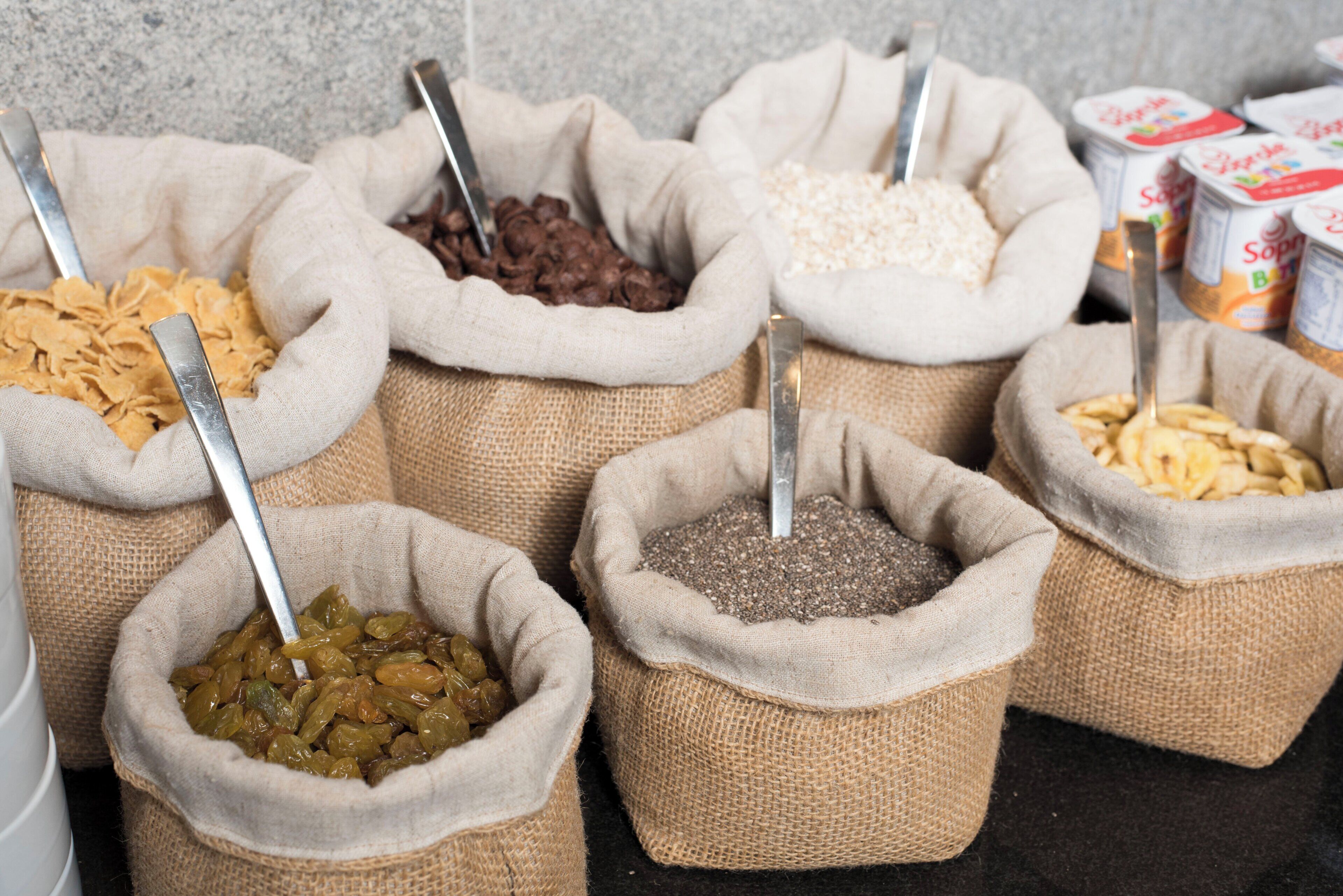 A variety of food items in burlap sacks with spoons, including what appears to be dried fruits, grains, and possibly legumes.