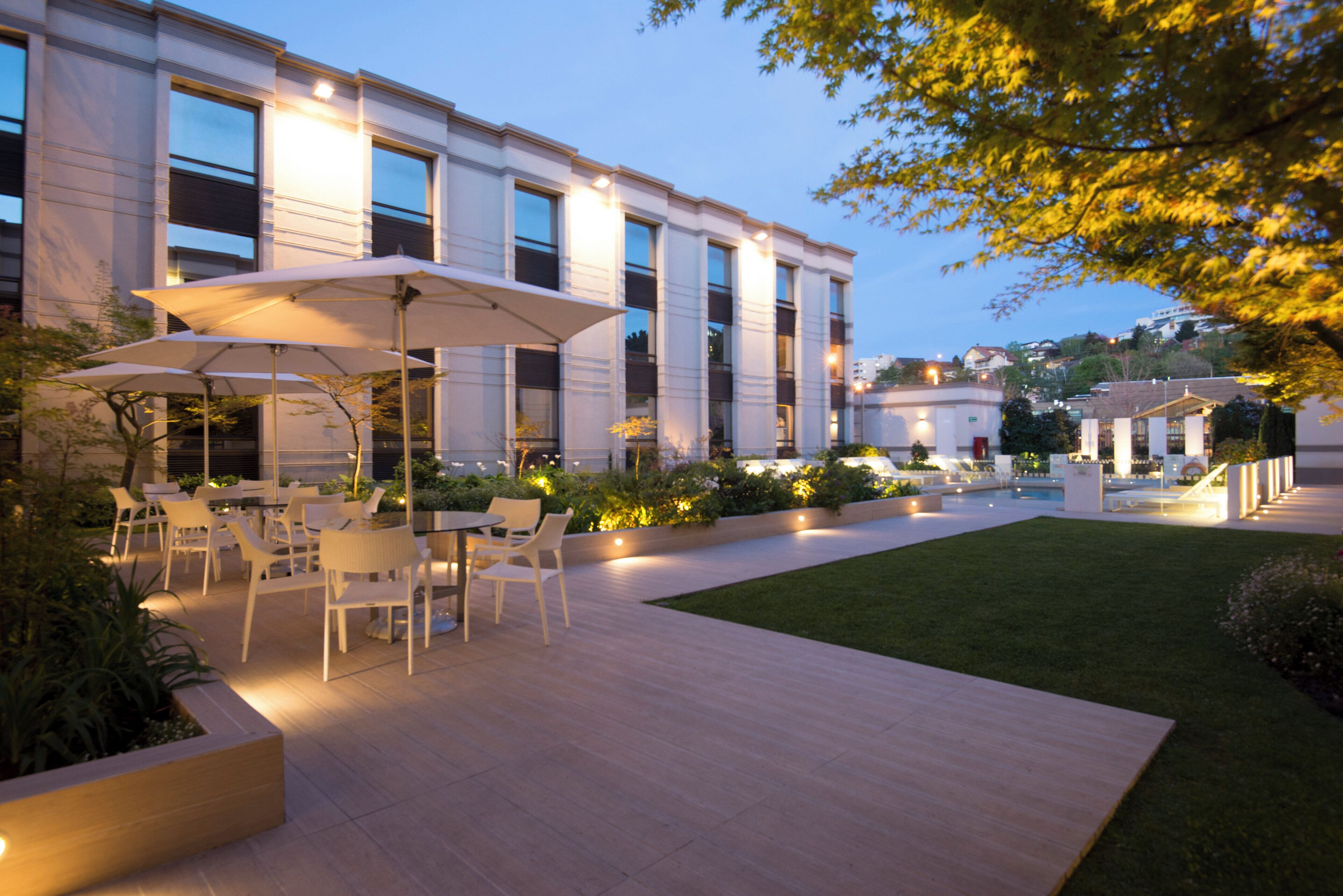 An outdoor patio area with tables and chairs, surrounded by a well-maintained lawn and a building with large windows.
