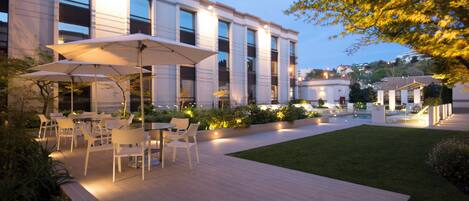 An outdoor patio area with tables and chairs, surrounded by a well-maintained lawn and a building with large windows.