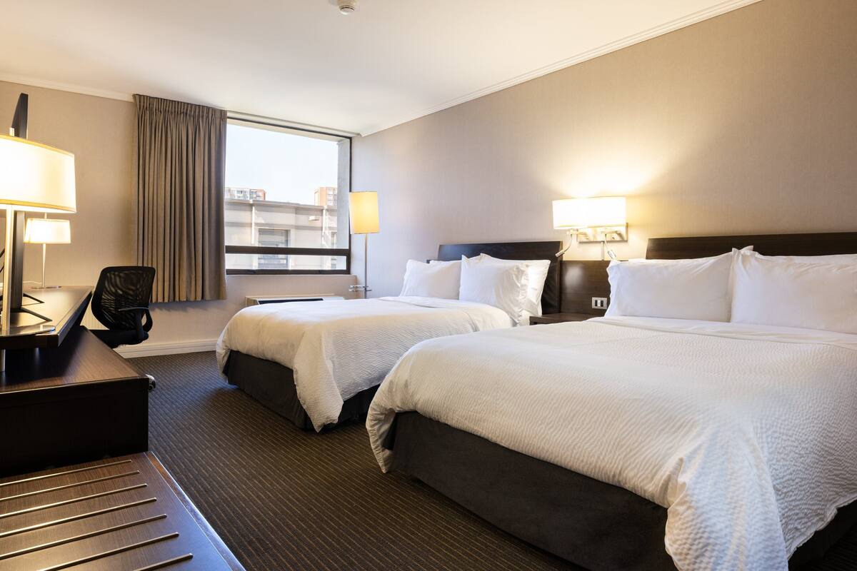 A hotel room with two beds, a desk, and a chair. There are two bedside lamps and a window with curtains.