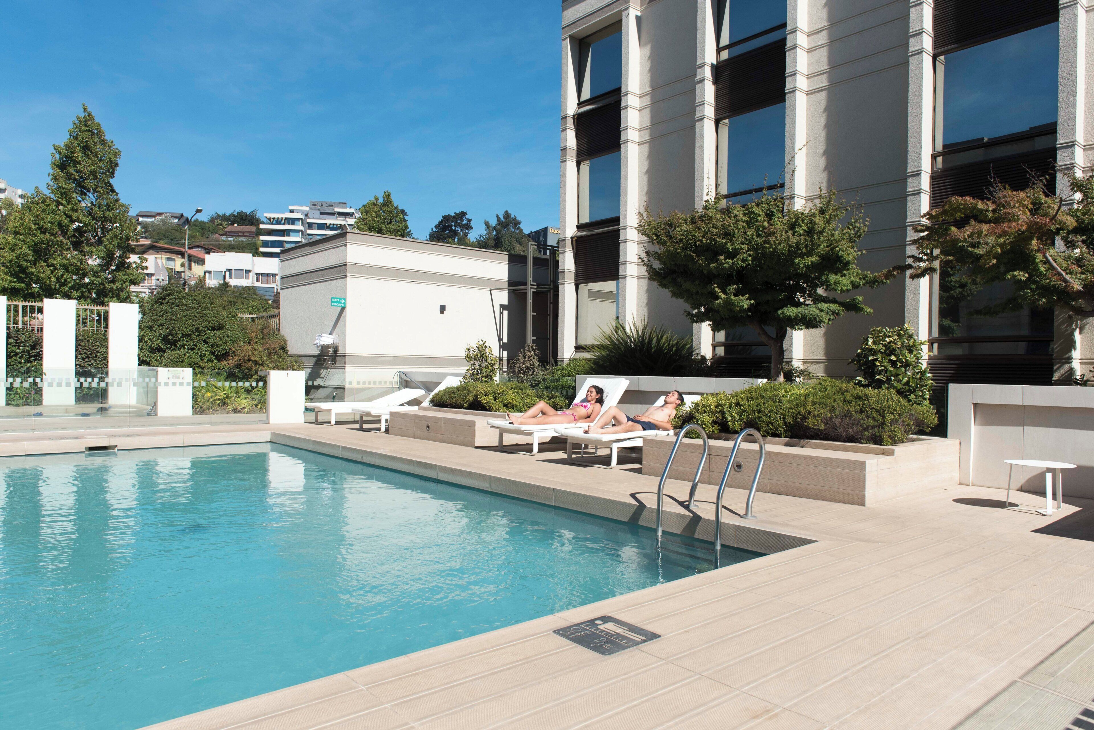 A swimming pool with lounge chairs and a small table, surrounded by greenery and a building with large windows.