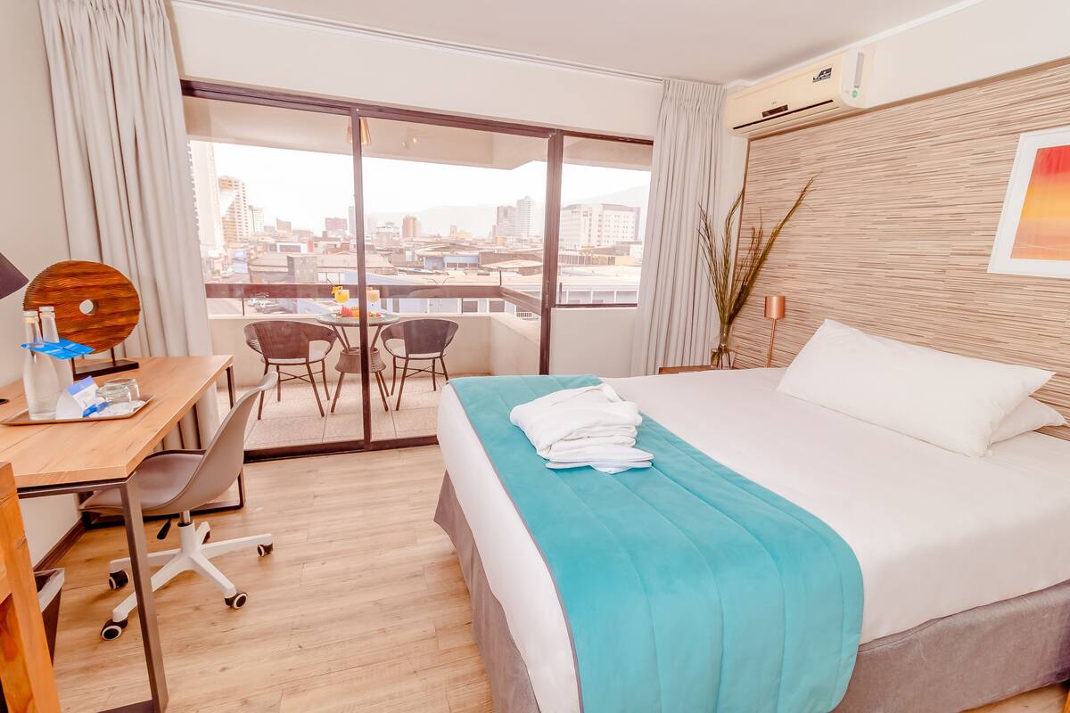 A hotel room with a bed, a desk with a chair, a small table, and a view of the city through large windows.