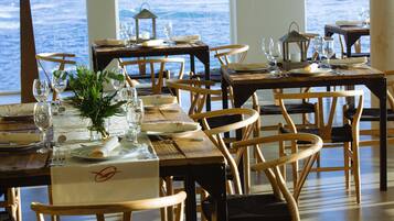 A restaurant with wooden tables and chairs, set against a large window overlooking the sea.