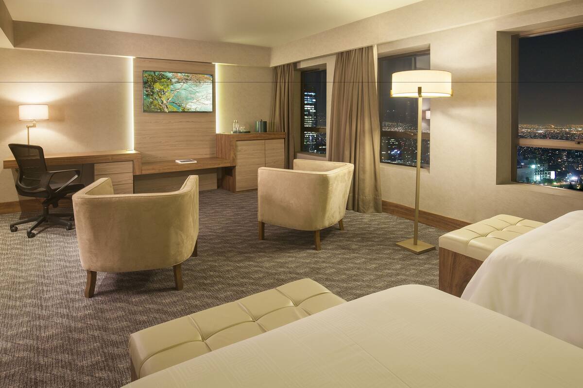 Hotelkamer met een bureau, twee fauteuils, een bed, een nachtkastje, een lamp, een schilderij en uitzicht op de stad.