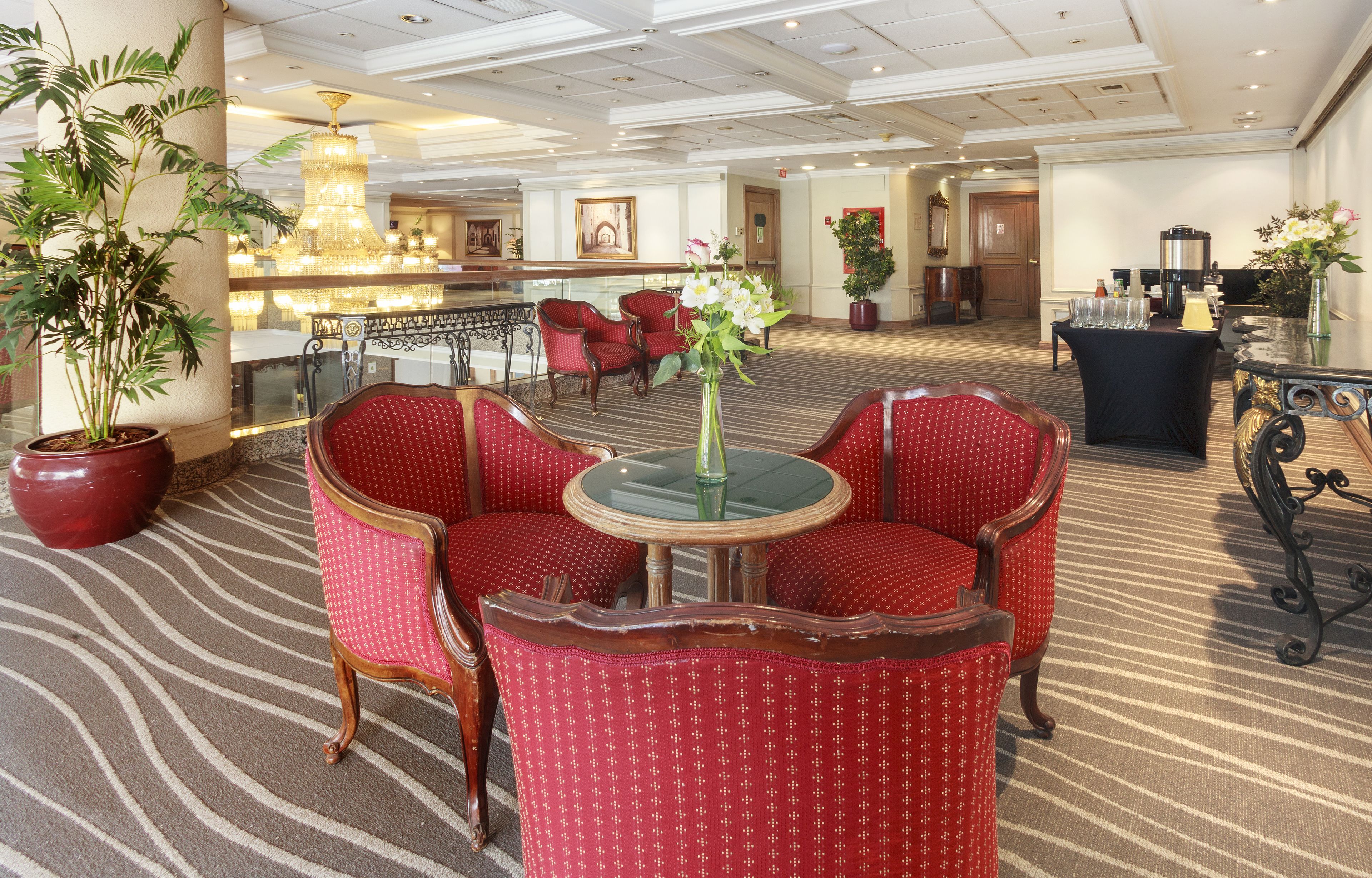 Un vestíbulo de hotel con sillones tapizados en rojo, una mesa redonda con un florero, una gran lámpara de araña y un mostrador de recepción.
