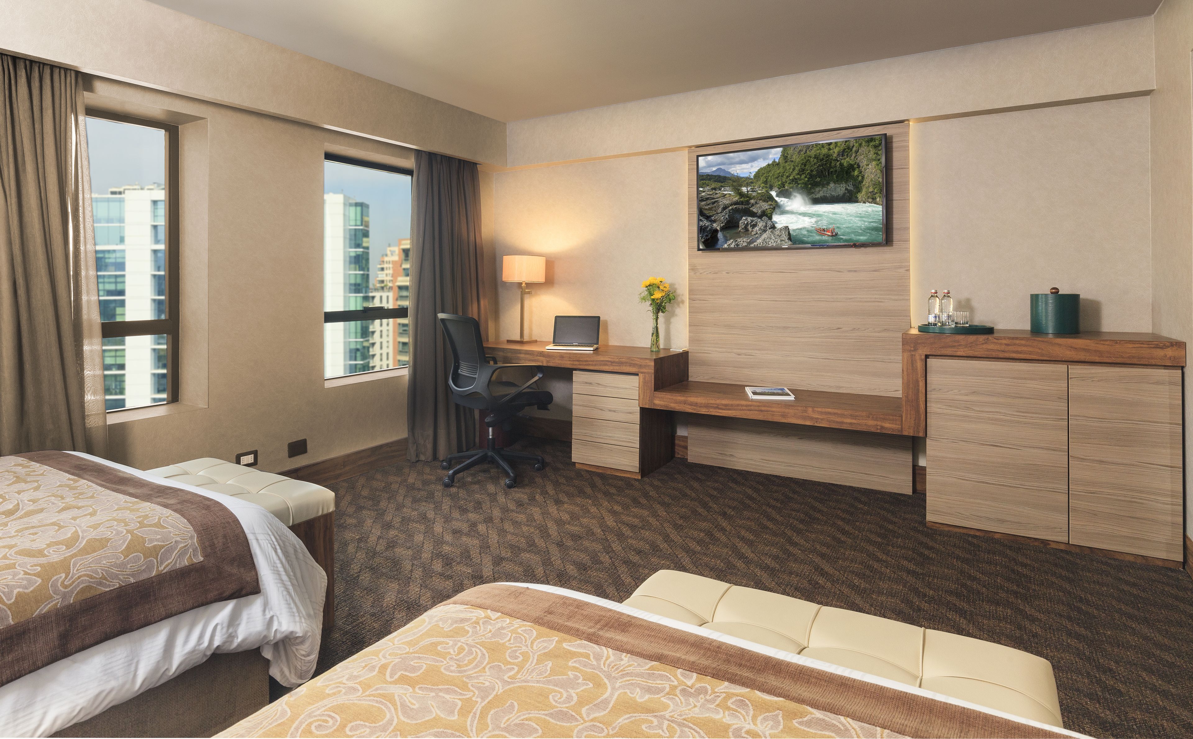 Habitación de hotel con dos camas, un escritorio con computadora, una silla, televisor en la pared y ventana con cortinas.