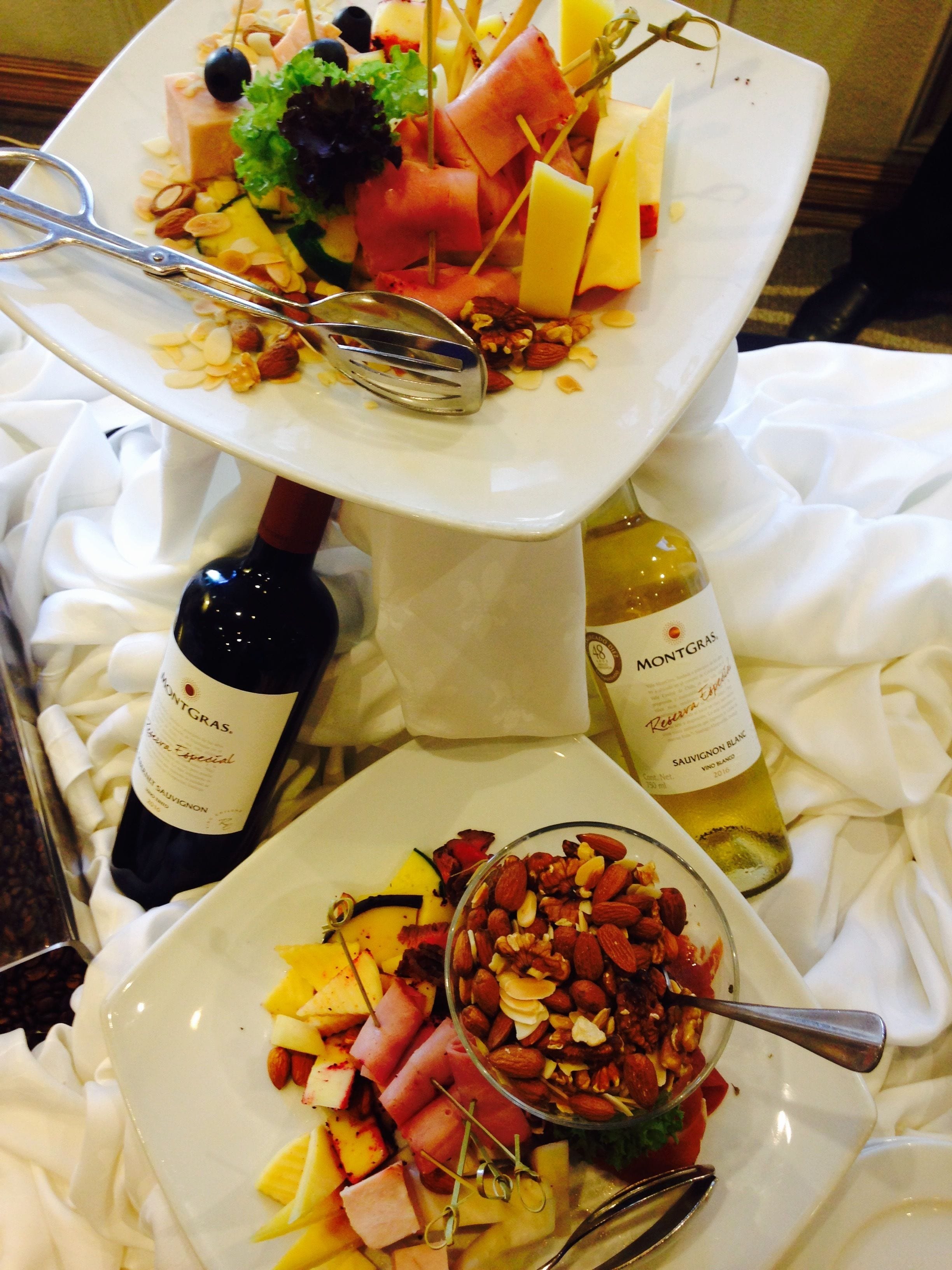 Ett serveringsfat i flera våningar med olika sorters ostar, frukter och nötter, tillsammans med två flaskor vin.
