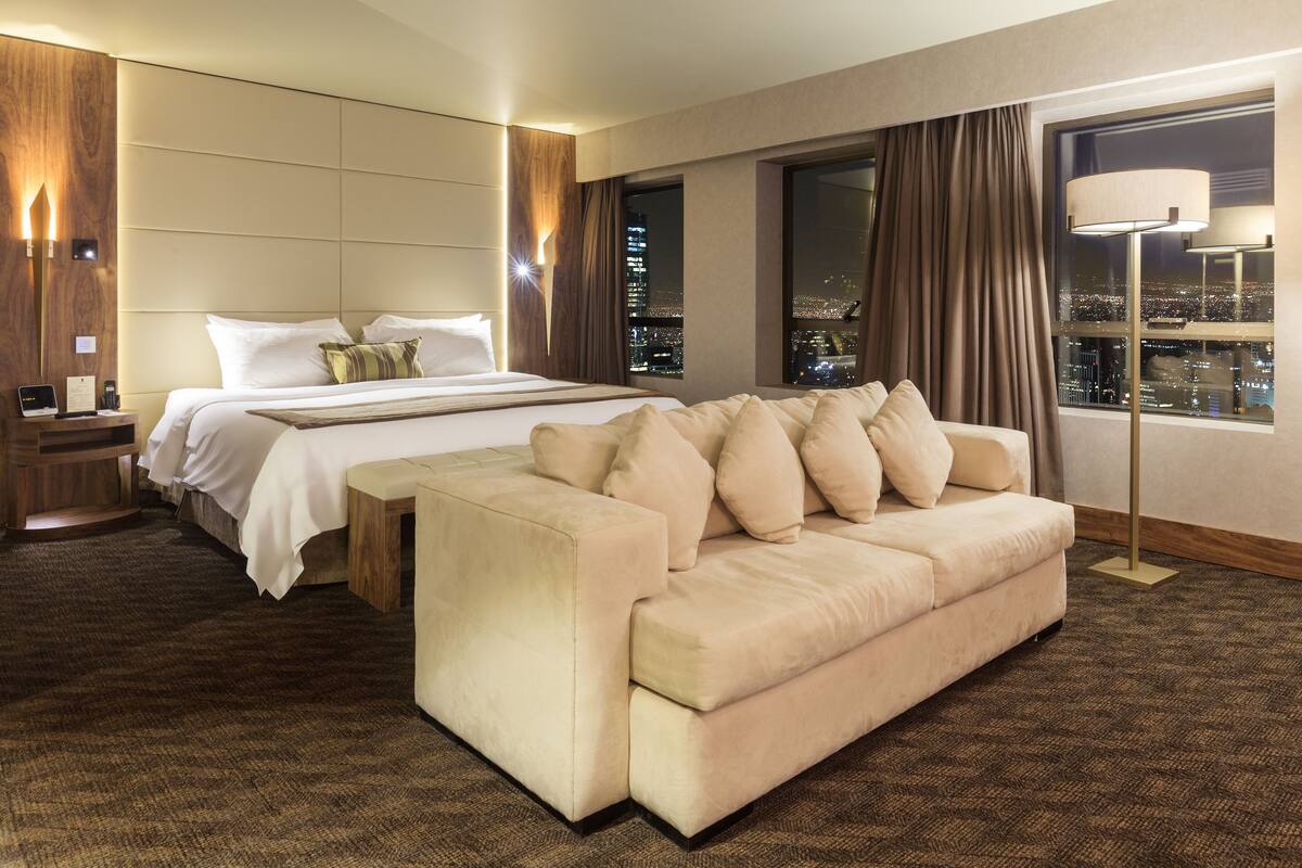 Een hotelkamer met een groot bed, een slaapbank, een nachtkastje en uitzicht op de stad 's nachts.