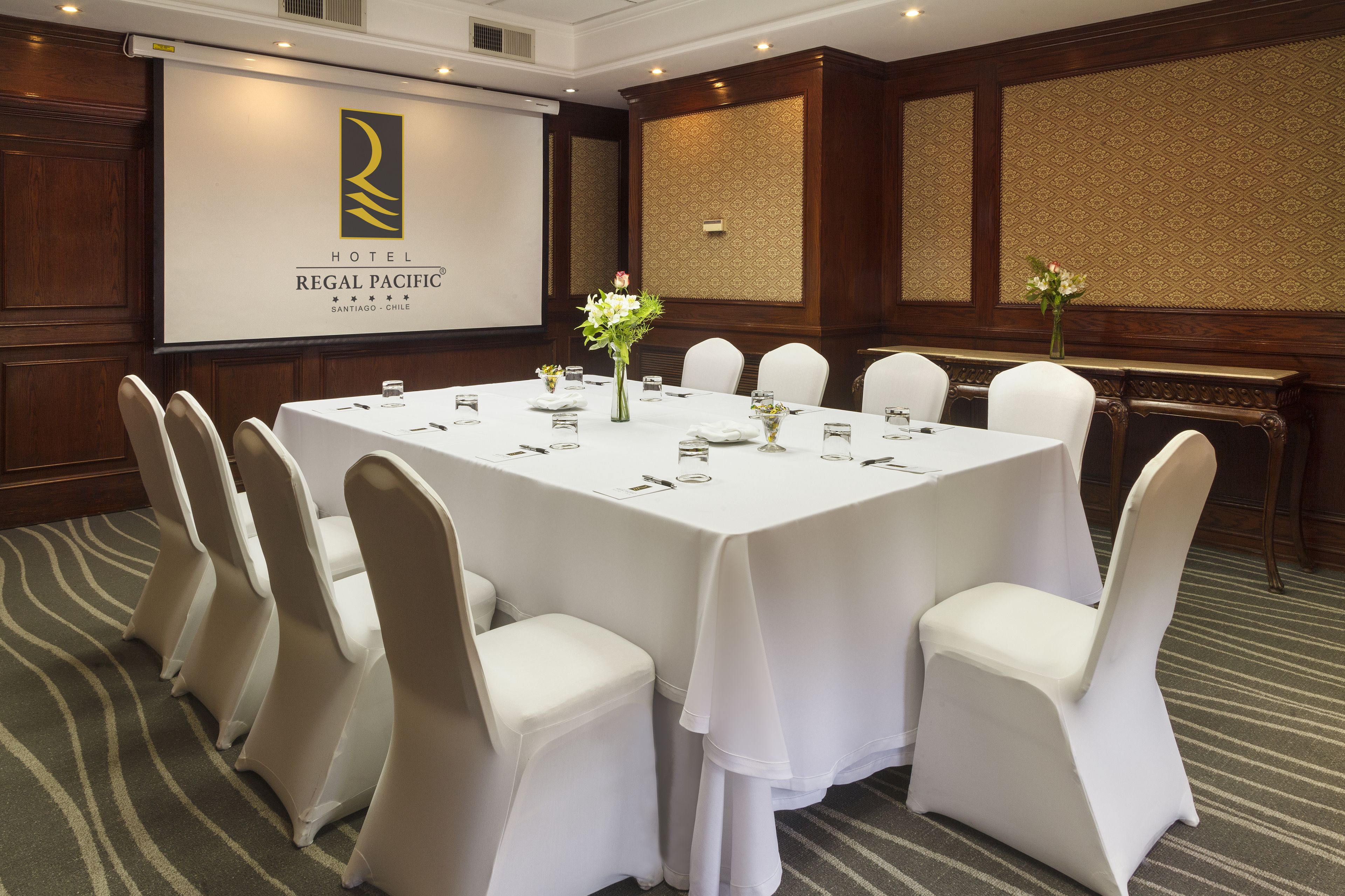 Un salón de conferencias con una mesa grande, sillas blancas y una pantalla que muestra el nombre del hotel.