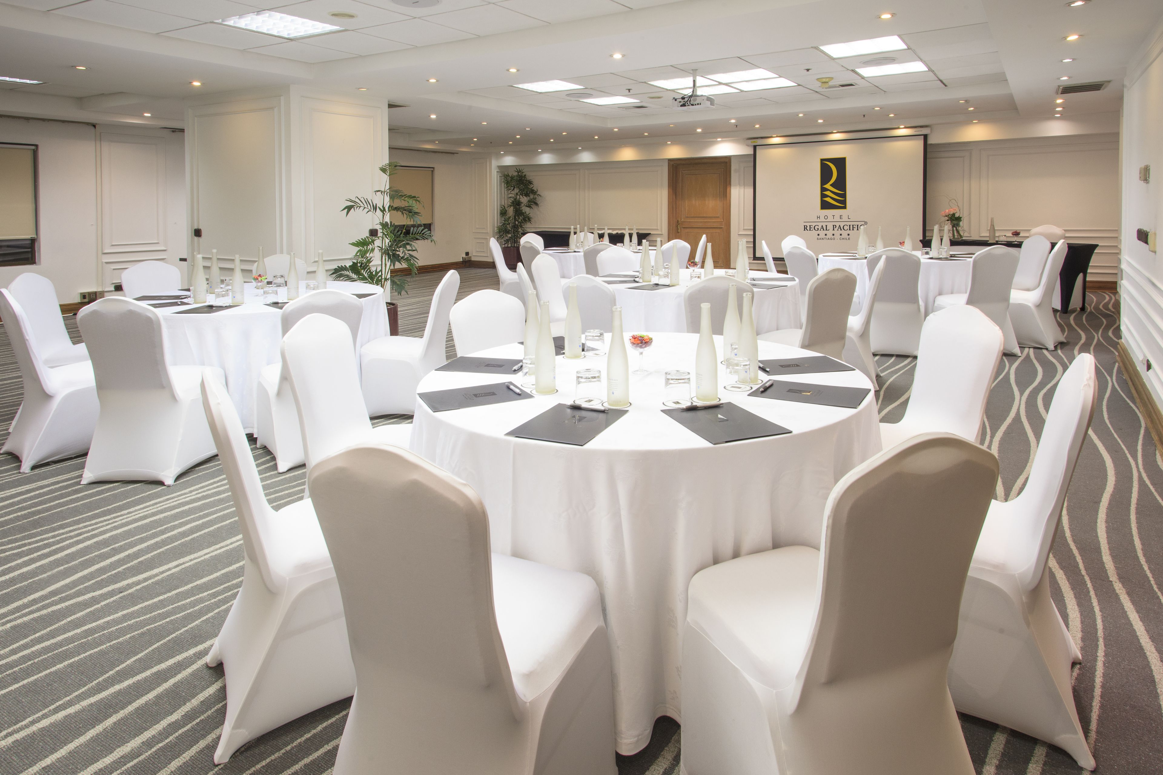 Un salón de conferencias con mesas redondas, sillas blancas y piso alfombrado.