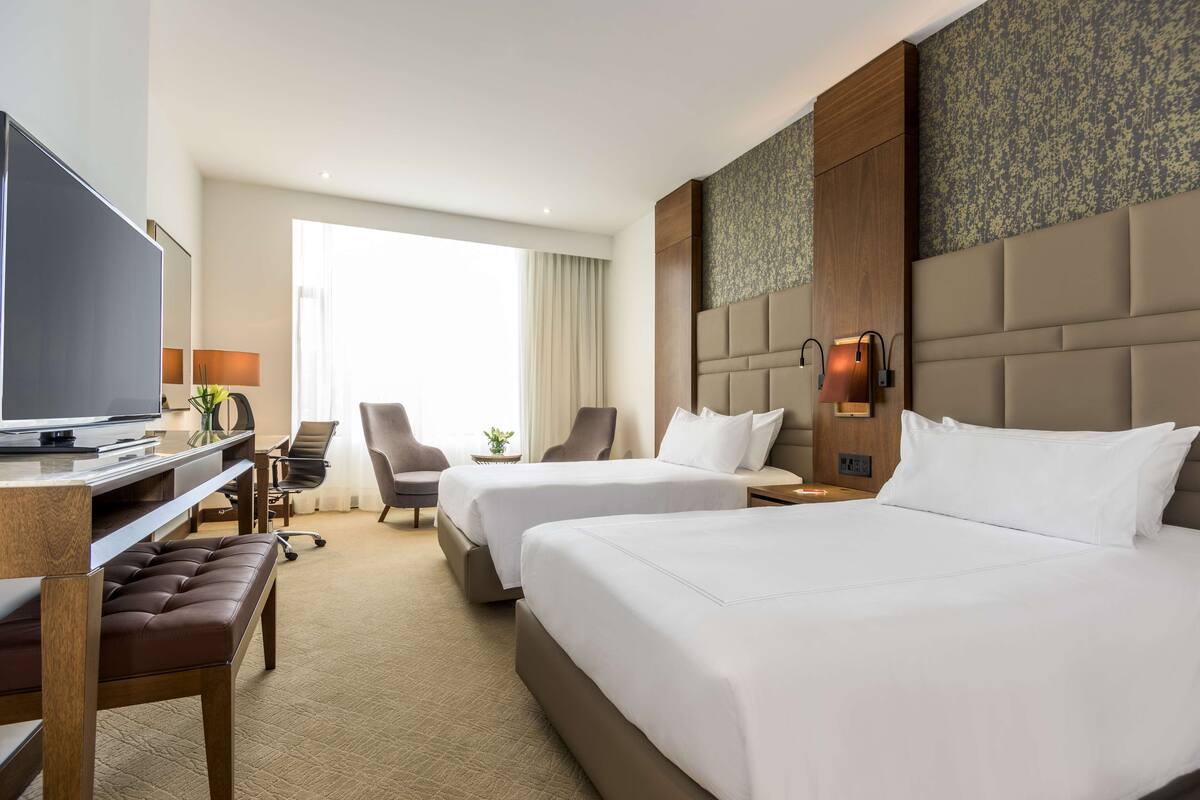 A hotel room with two beds, a desk with a TV, a chair, and a small table with a plant.