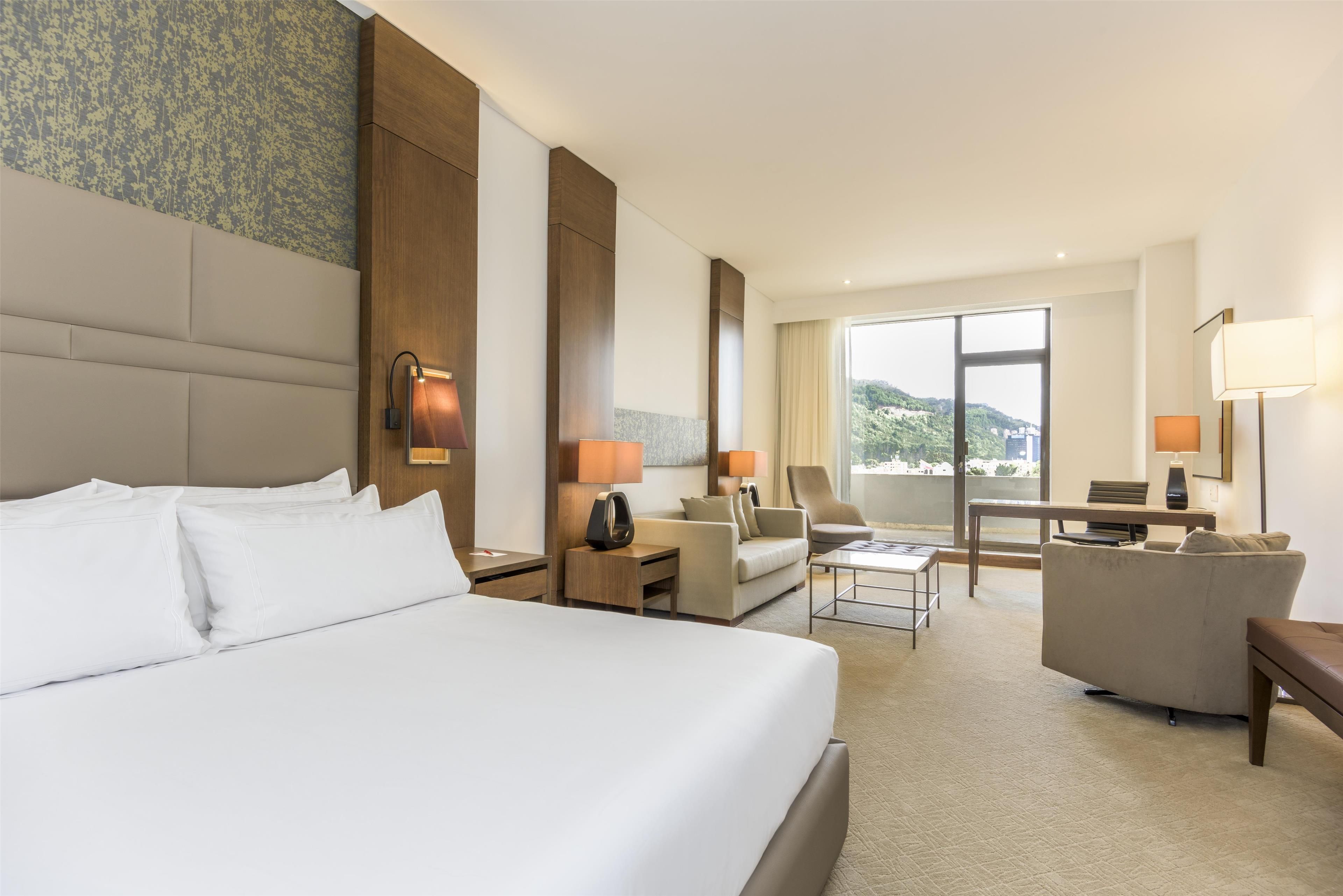A hotel room with a large bed, a sofa, a desk, and a chair. There is a window with a view of greenery.