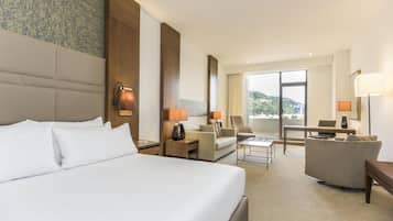 A hotel room with a large bed, a sofa, a desk, and a chair. There is a window with a view of greenery.