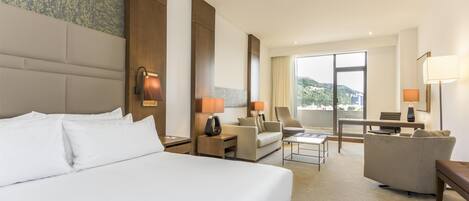 A hotel room with a large bed, a sofa, a desk, and a chair. There is a window with a view of greenery.