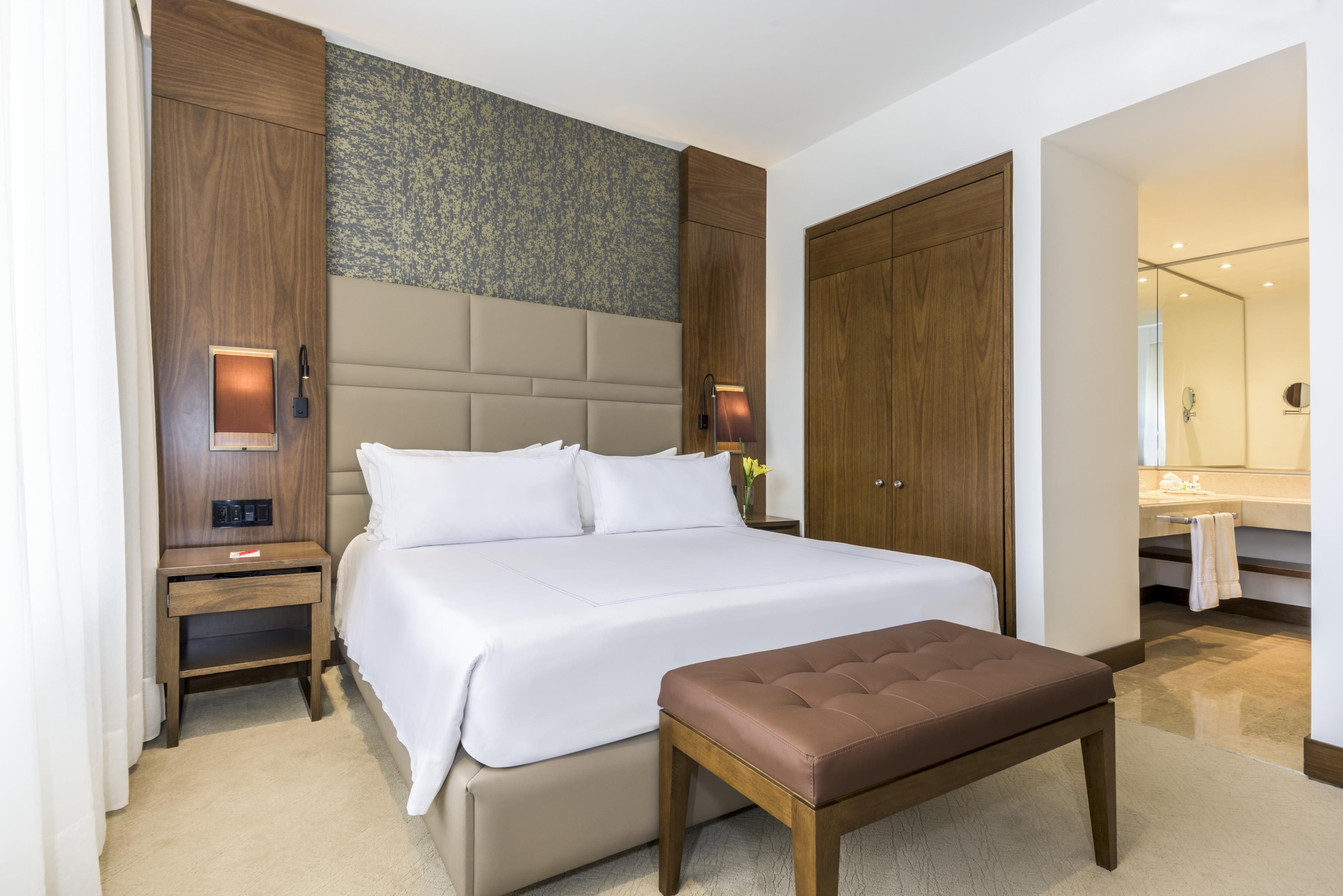 A modern hotel room with a large bed, wooden headboard, bedside lamps, a small table, and a bench.