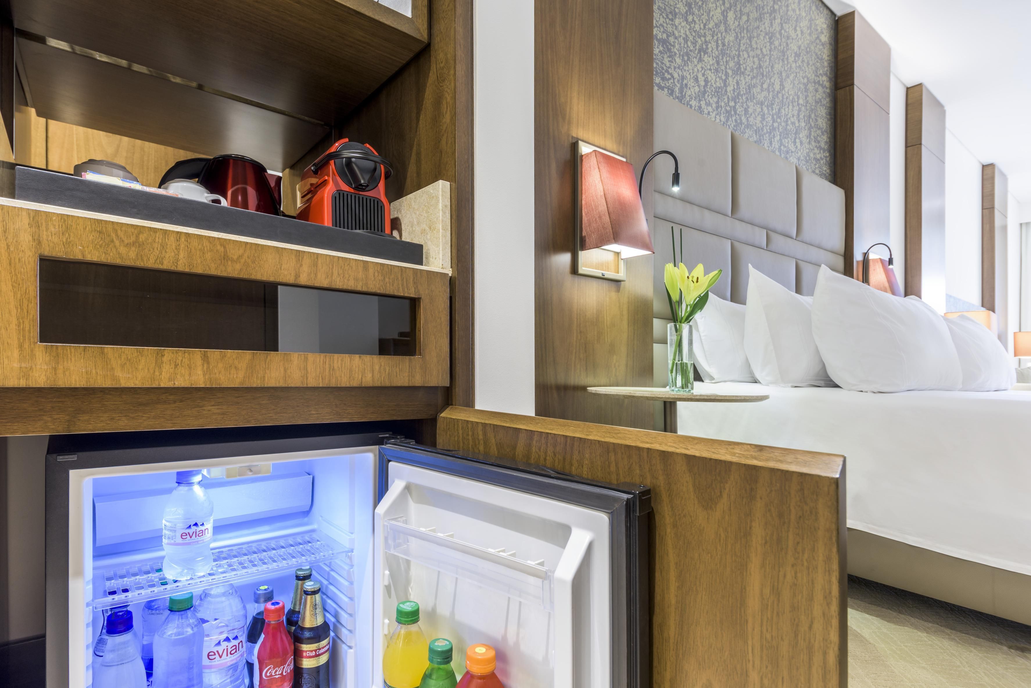 A hotel room with a built-in refrigerator stocked with beverages, a bedside table with a lamp, and a bed with white linens.