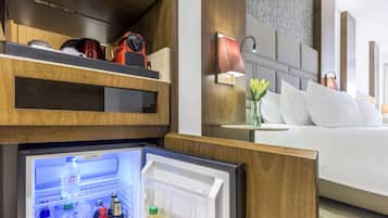 A hotel room with a built-in refrigerator stocked with beverages, a bedside table with a lamp, and a bed with white linens.